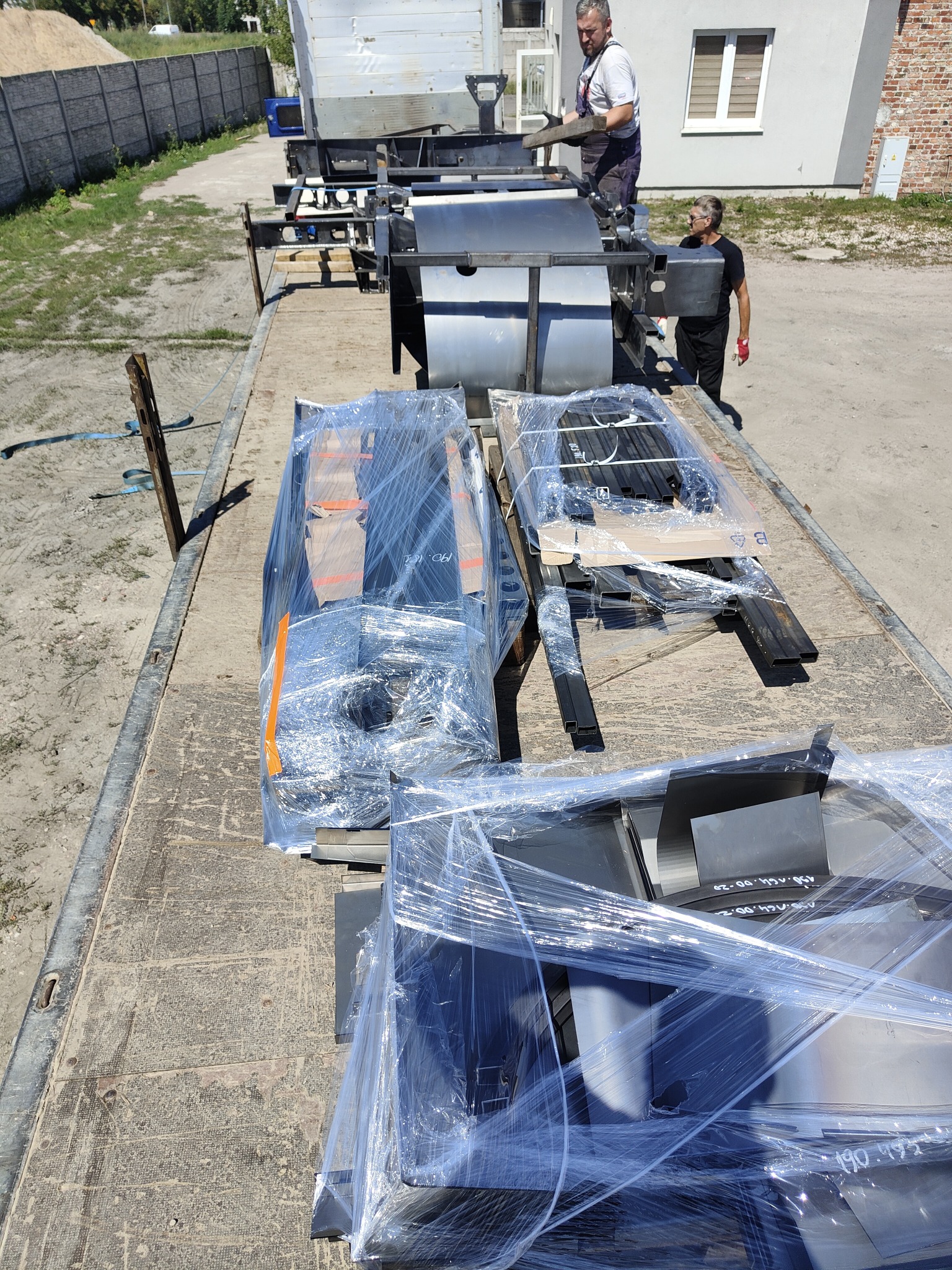 Transport elementów konstrukcji stalowej na platformie ciężarówki, pracownicy w tle, elementy zabezpieczone folią stretch.