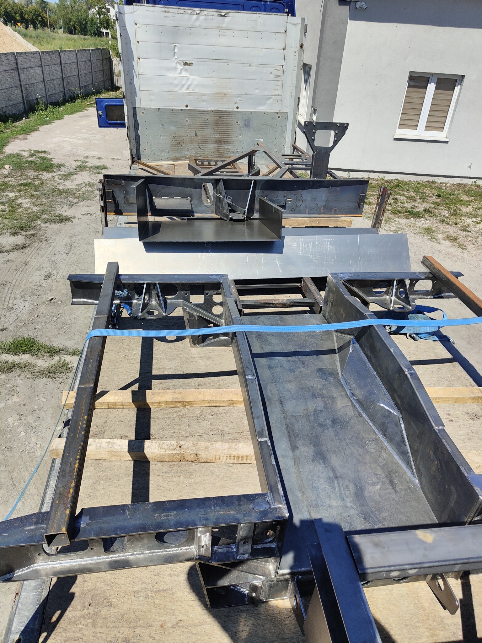 Elementy konstrukcji stalowej, prawdopodobnie ramy pojazdu, transportowane na platformie ciężarówki, zabezpieczone niebieskim pasem, w tle budynek i ogrodzenie.