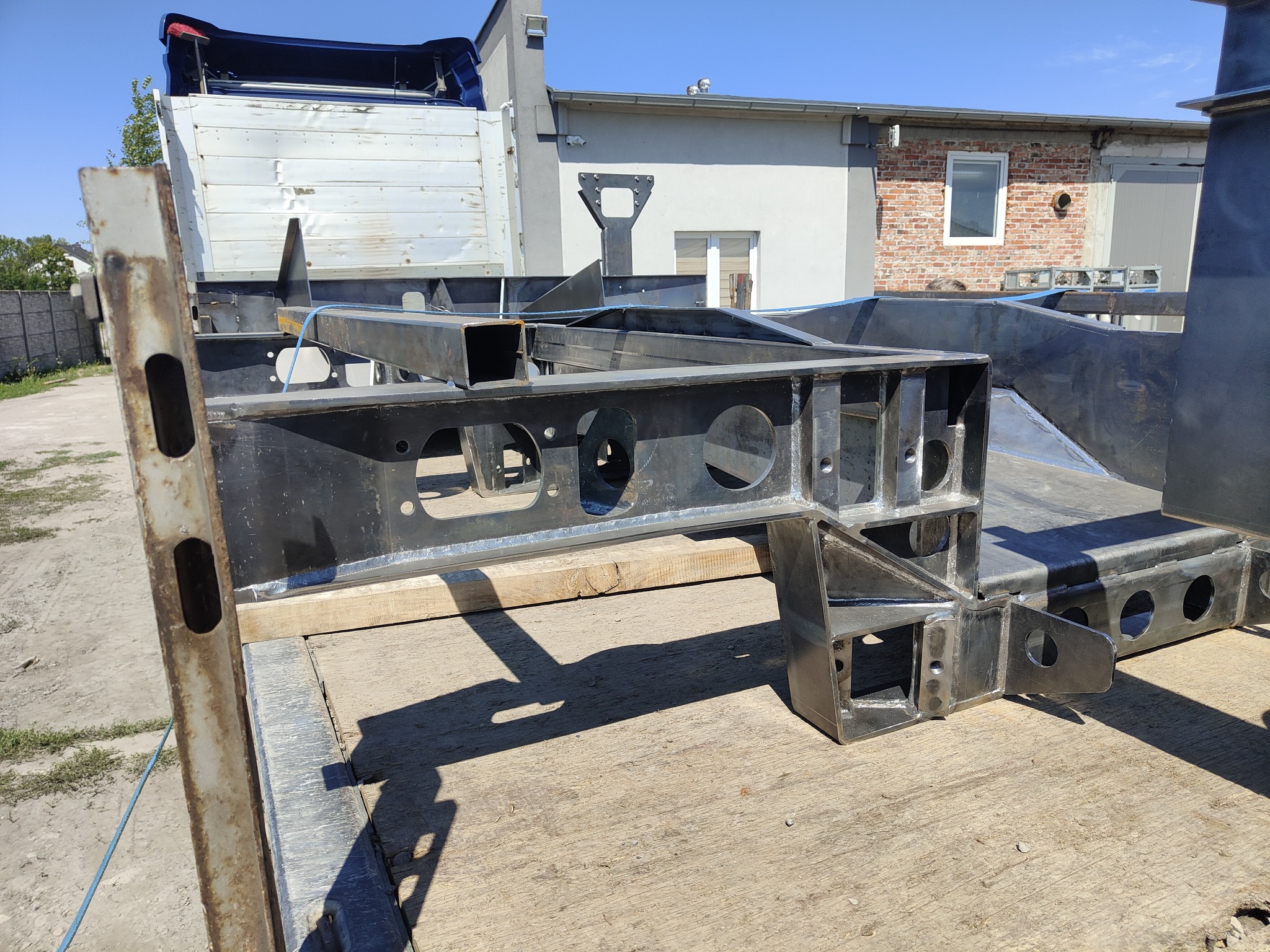 Surowa, czarna konstrukcja stalowa z profili zamkniętych, z licznymi otworami, leżąca na drewnianej platformie przyczepy samochodowej, na tle budynku z ceglaną ścianą i błękitnego nieba.
