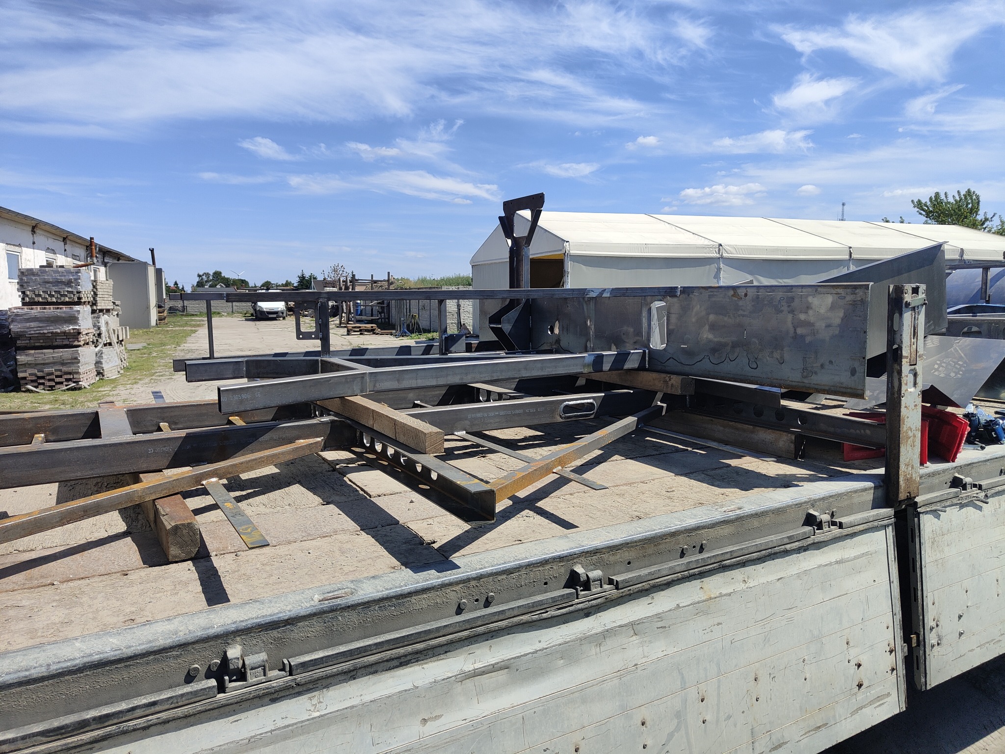 Elementy konstrukcji stalowej, prawdopodobnie hali, ułożone na przyczepie samochodu ciężarowego, w tle budynek i namiot magazynowy.