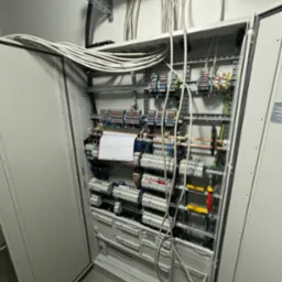 Otwarta szafa rozdzielcza z licznymi bezpiecznikami, przewodami i listwami zaciskowymi, widoczne kable prowadzone w korytkach na suficie.