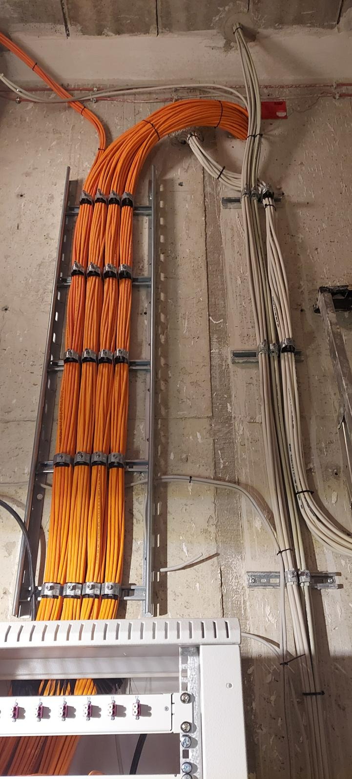 Pomarańczowe i białe kable elektryczne uporządkowane w korytkach i przymocowane do surowej ściany, widoczne elementy szafy sterowniczej na dole kadru.