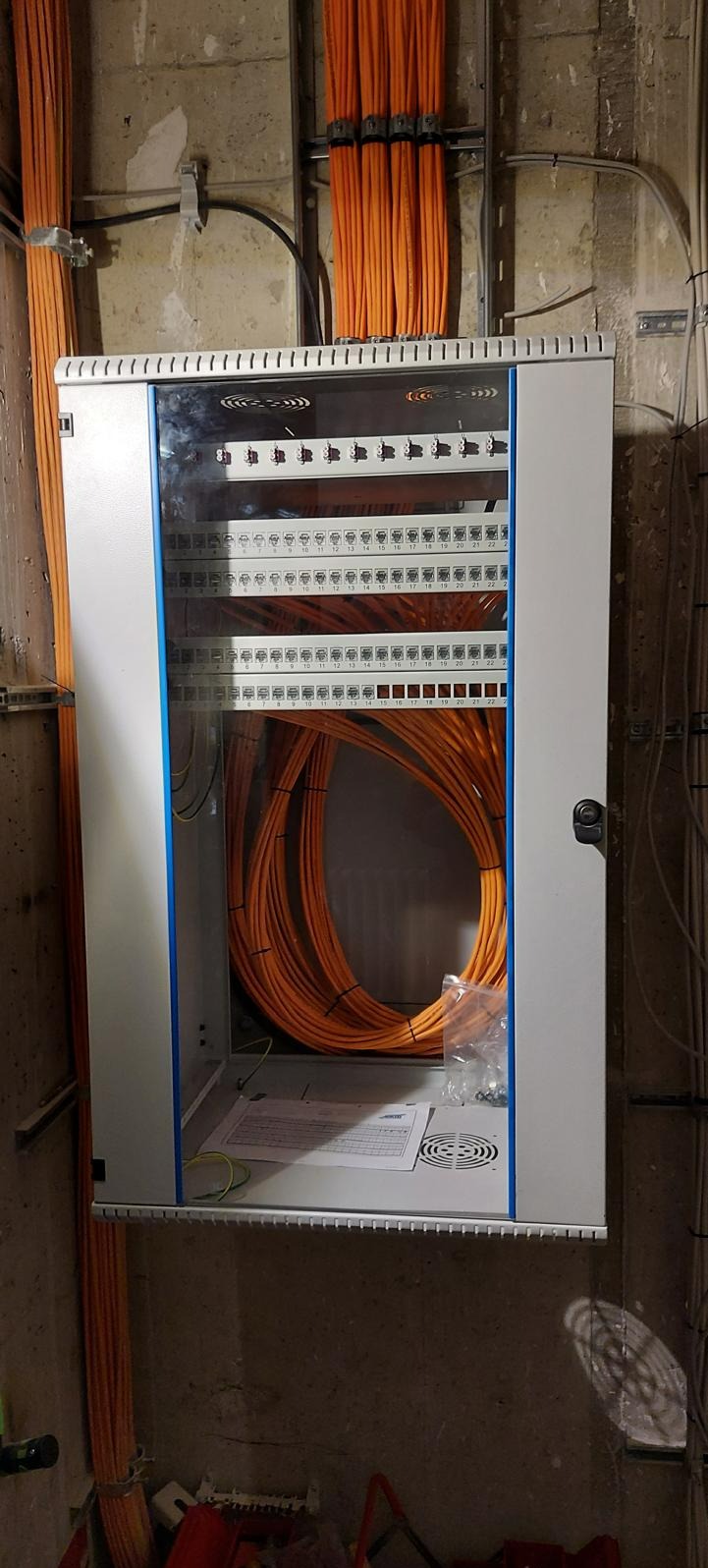 Otwarta szafa rack z pomarańczowymi kablami sieciowymi wewnątrz, zamontowana na surowej ścianie z widocznymi instalacjami.