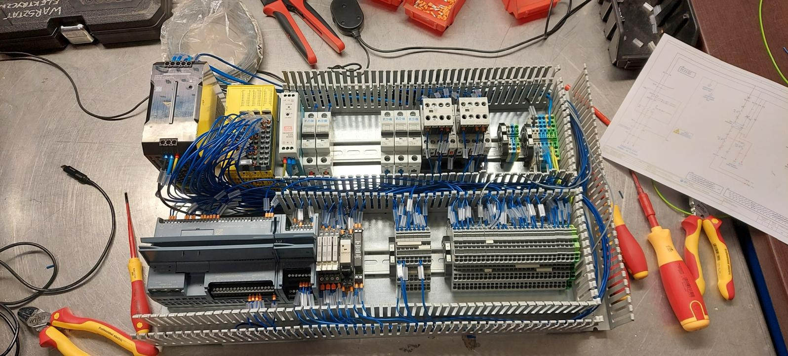 Rozdzielnica elektryczna w trakcie montażu, widoczne przewody, bezpieczniki, sterowniki PLC Siemens, schemat elektryczny i narzędzia: wkrętaki i szczypce.