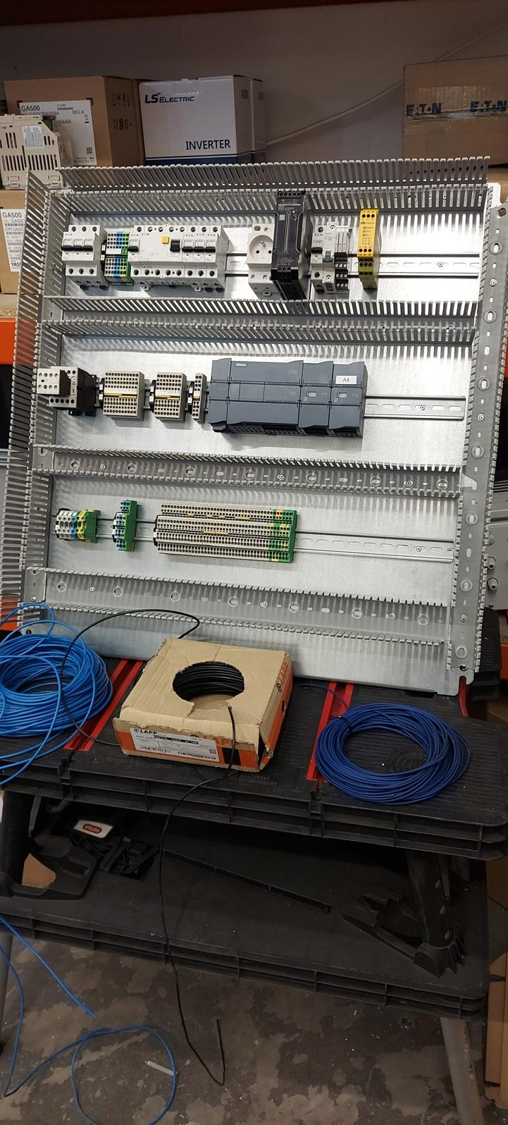 Metalowa szafa sterownicza w trakcie montażu, widoczne moduły, bezpieczniki, złączki, przewody niebieskie i czarne w kartonie marki LAPP, w tle kartony z elektroniką.