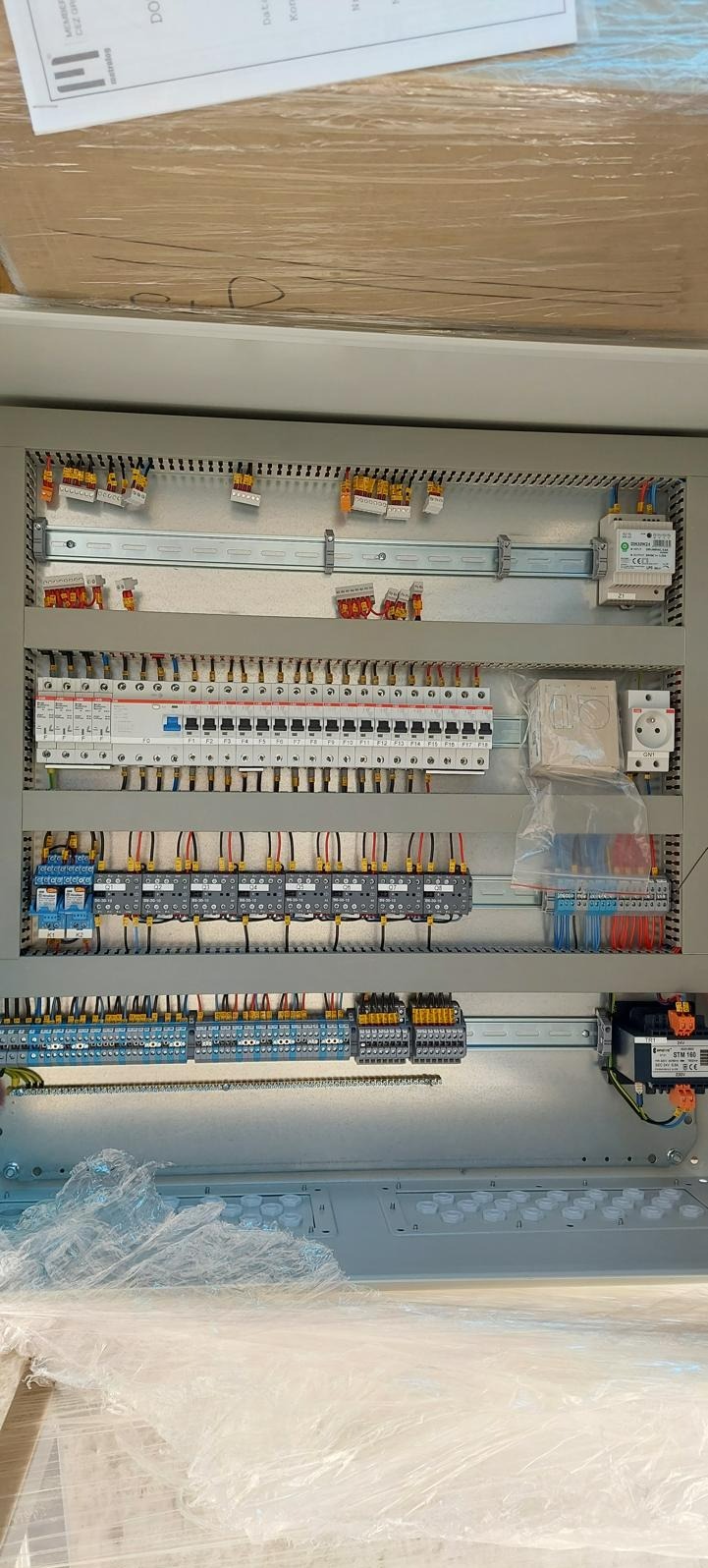 Wnętrze szarej szafy rozdzielczej z zamontowanymi bezpiecznikami, stycznikami i przewodami, widoczne listwy DIN oraz gniazdo elektryczne w folii ochronnej.