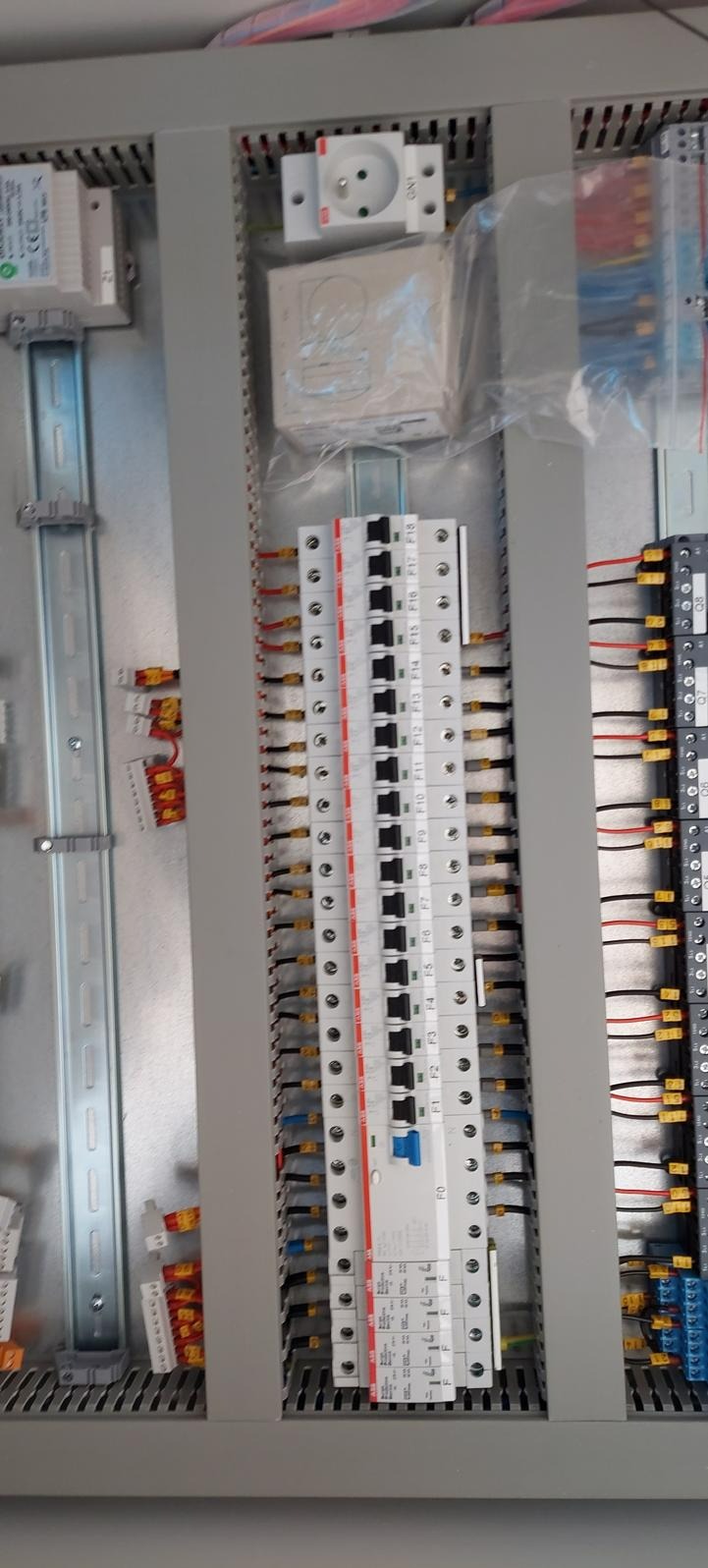 Wnętrze szafy sterowniczej z widocznymi bezpiecznikami, listwami zaciskowymi i przewodami o różnych kolorach, gniazdo elektryczne w obudowie, częściowo zapakowane elementy.