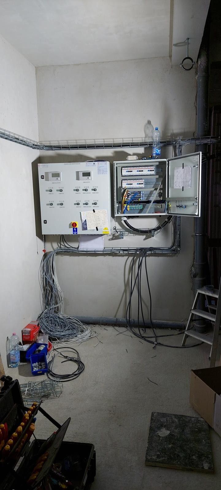 Rozdzielnica elektryczna z otwartą skrzynką bezpieczników, widoczne kable, narzędzia i akcesoria elektryczne w surowym pomieszczeniu.