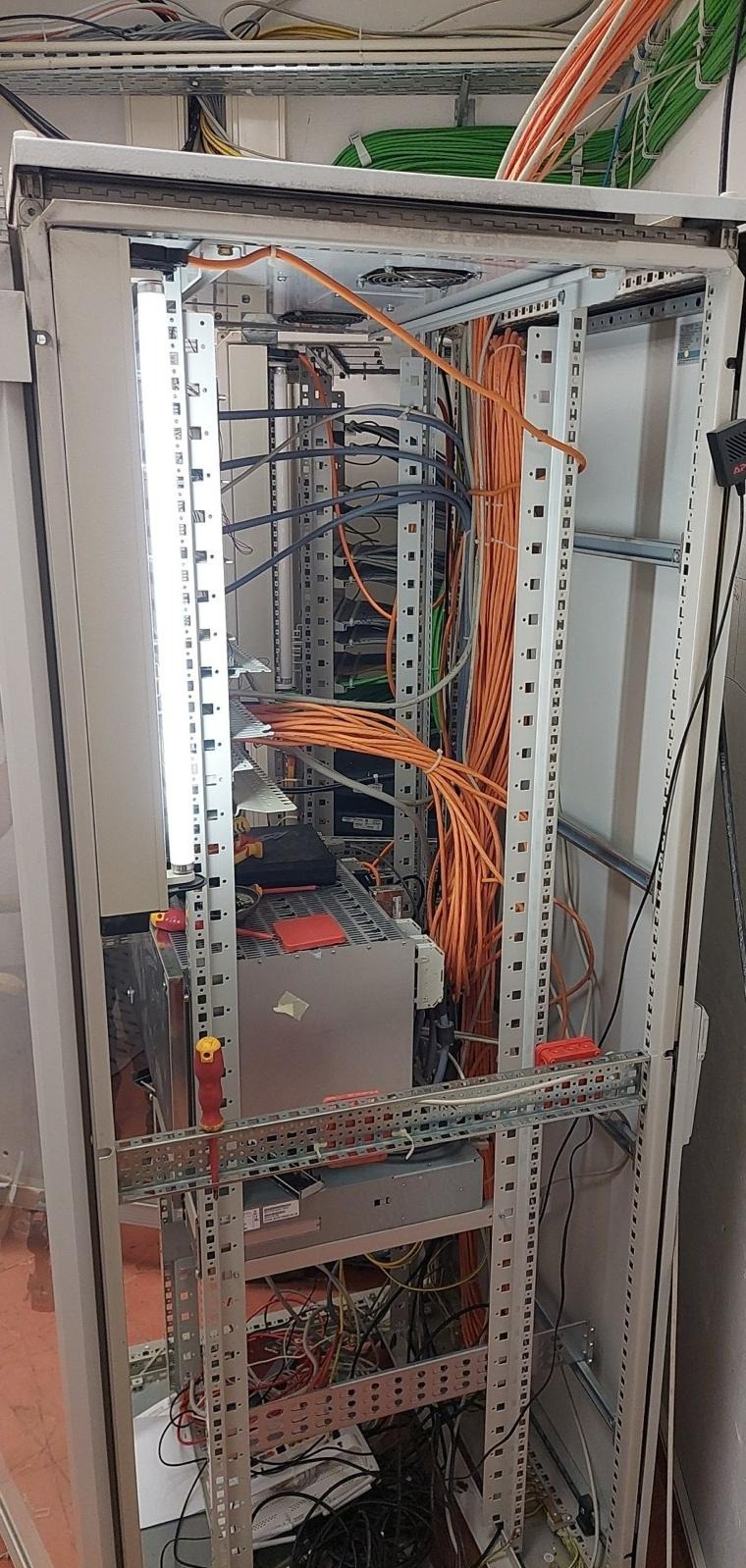 Otwarta szafa serwerowa z licznymi kablami pomarańczowymi i szarymi, widoczne urządzenia, listwa oświetleniowa LED po lewej stronie, narzędzia na półkach, oraz przewody wychodzące z góry szafy.