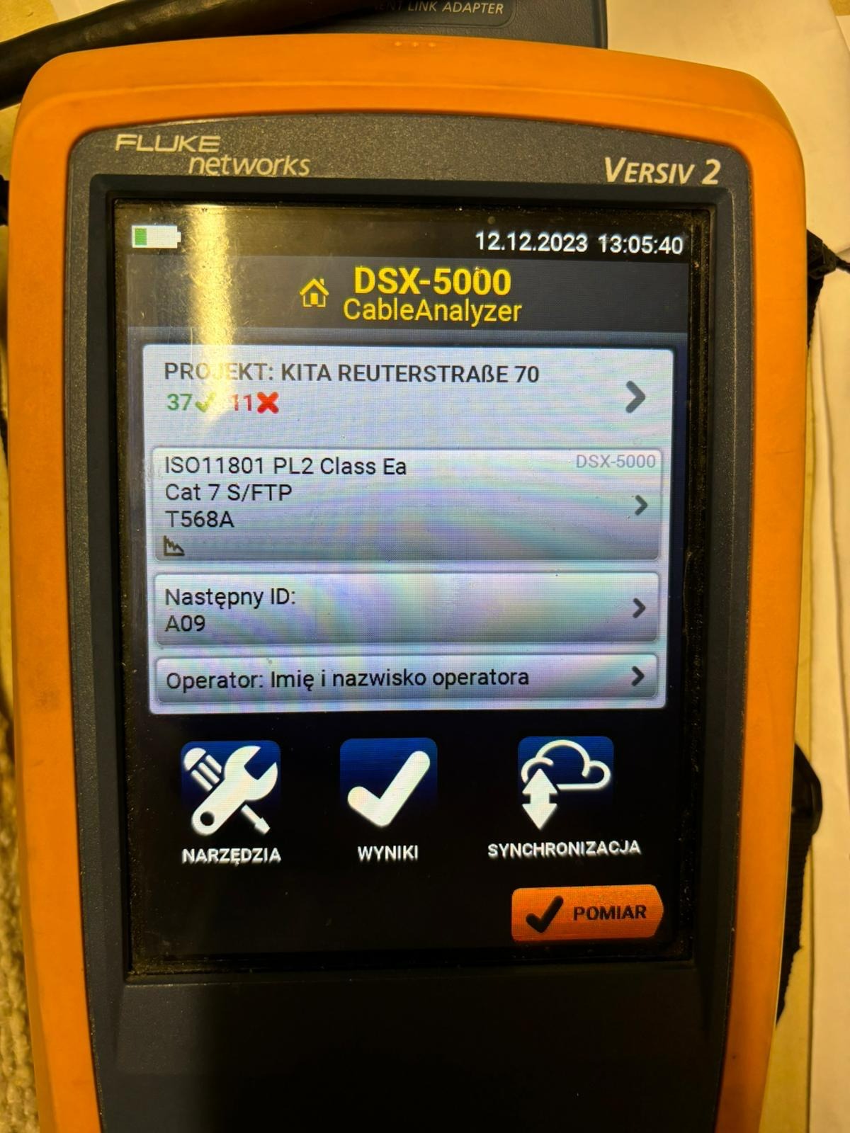 Pomarańczowy tester kabli Fluke Networks Versiv 2 DSX-5000 CableAnalyzer wyświetlający wyniki pomiaru okablowania strukturalnego, w tym informacje o projekcie, standardzie ISO11801, kategorii Cat 7...