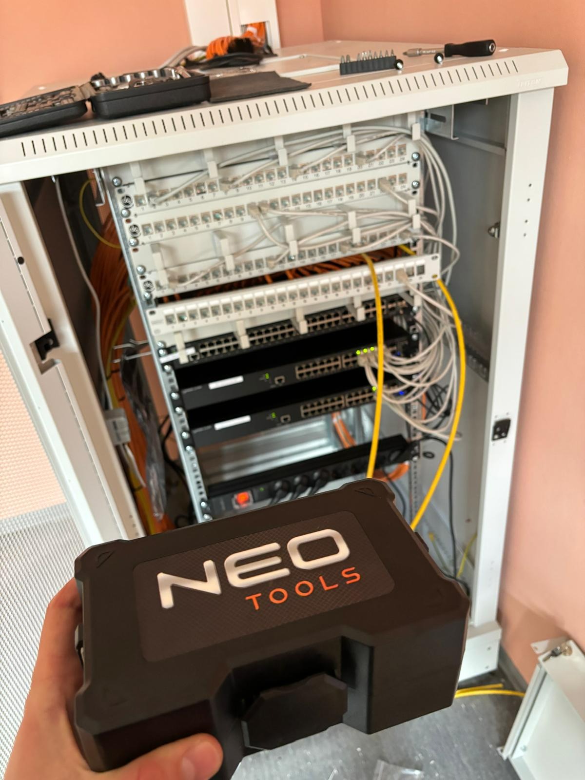 Szafa rack z patch panelem, switchem i okablowaniem, widziana z perspektywy osoby trzymającej skrzynkę narzędziową Neo Tools.