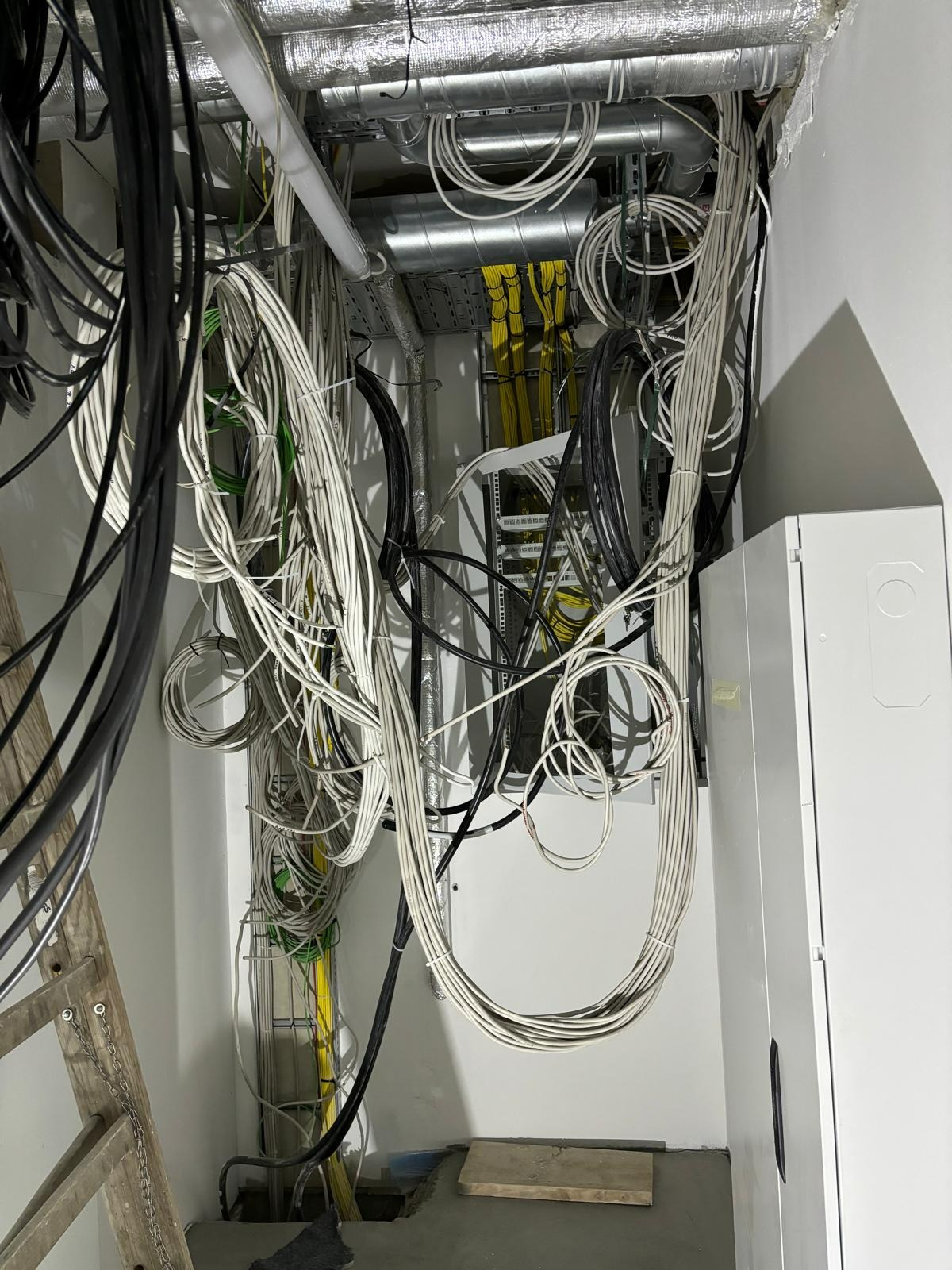 Skomplikowany układ przewodów elektrycznych w przestrzeni pod sufitem, widoczne rury wentylacyjne i drabina oparta o ścianę, częściowo zamaskowany panel elektryczny z żółtymi kablami w tle.