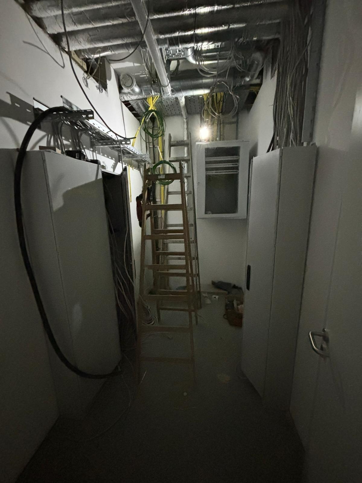 Wnętrze w trakcie prac elektrycznych: drabina oparta o ścianę z zamontowaną skrzynką elektryczną, widoczne kable i przewody pod sufitem oraz szafy elektryczne po bokach, oświetlenie robocze.