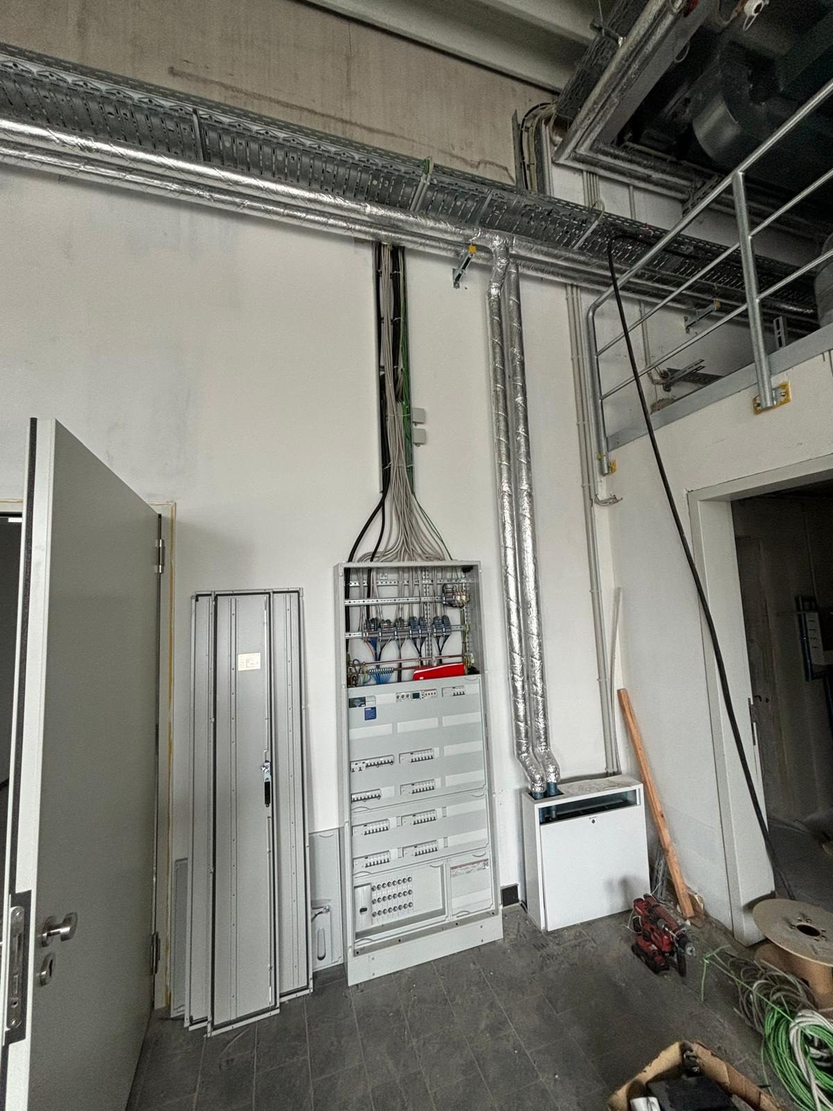 Otwarta szafa sterownicza z widocznymi kablami i bezpiecznikami w pomieszczeniu przemysłowym, z przewodami w metalowych korytach pod sufitem, obok biała obudowa urządzenia z izolowanymi rurami.