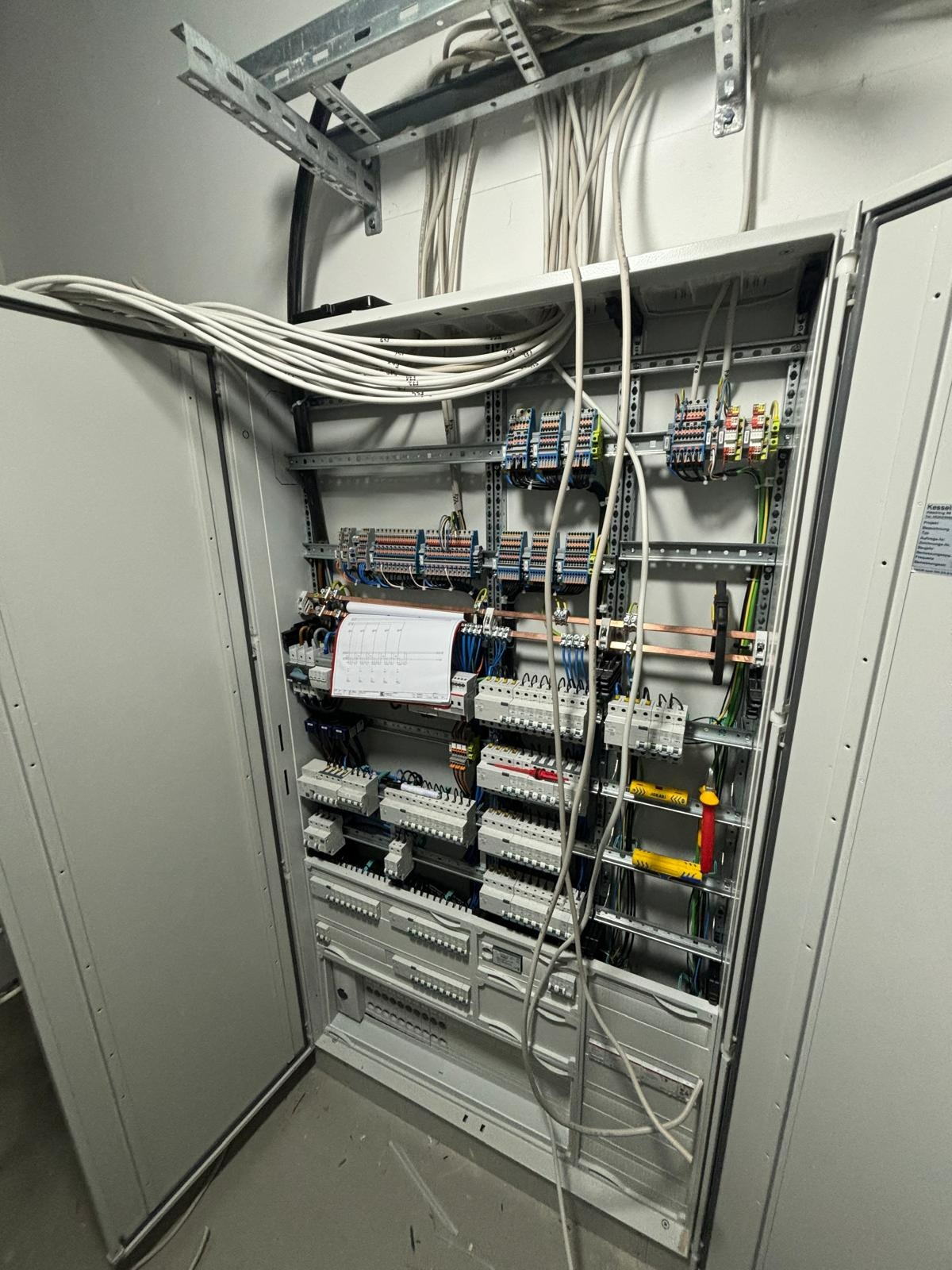 Otwarta szafa rozdzielcza z licznymi bezpiecznikami, przewodami i listwami zaciskowymi, widoczne kable prowadzone w korytkach na suficie.