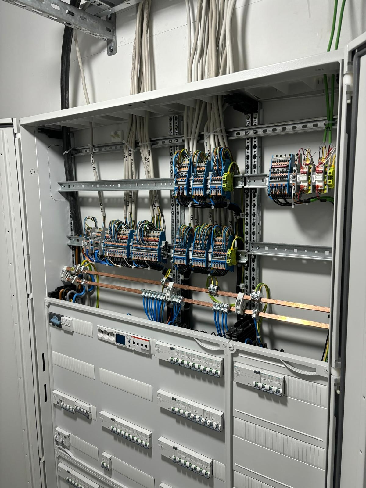 Otwarta szafa elektryczna z widocznymi bezpiecznikami, listwami zaciskowymi i okablowaniem, uporządkowana instalacja w szarej obudowie.