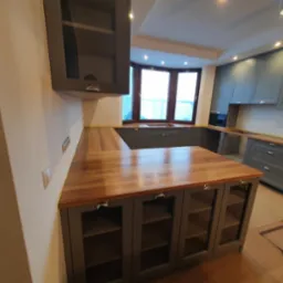 Szara zabudowa kuchenna z drewnianym blatem, widok na narożne okno i oświetlenie punktowe sufitowe, dolne szafki z przeszklonymi frontami.