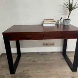 Biurko z ciemnego drewna z czarnymi, metalowymi nogami, z dwiema szufladami, na blacie książki, roślina doniczkowa i słoik z długopisami, na tle białej ściany z gniazdkami.