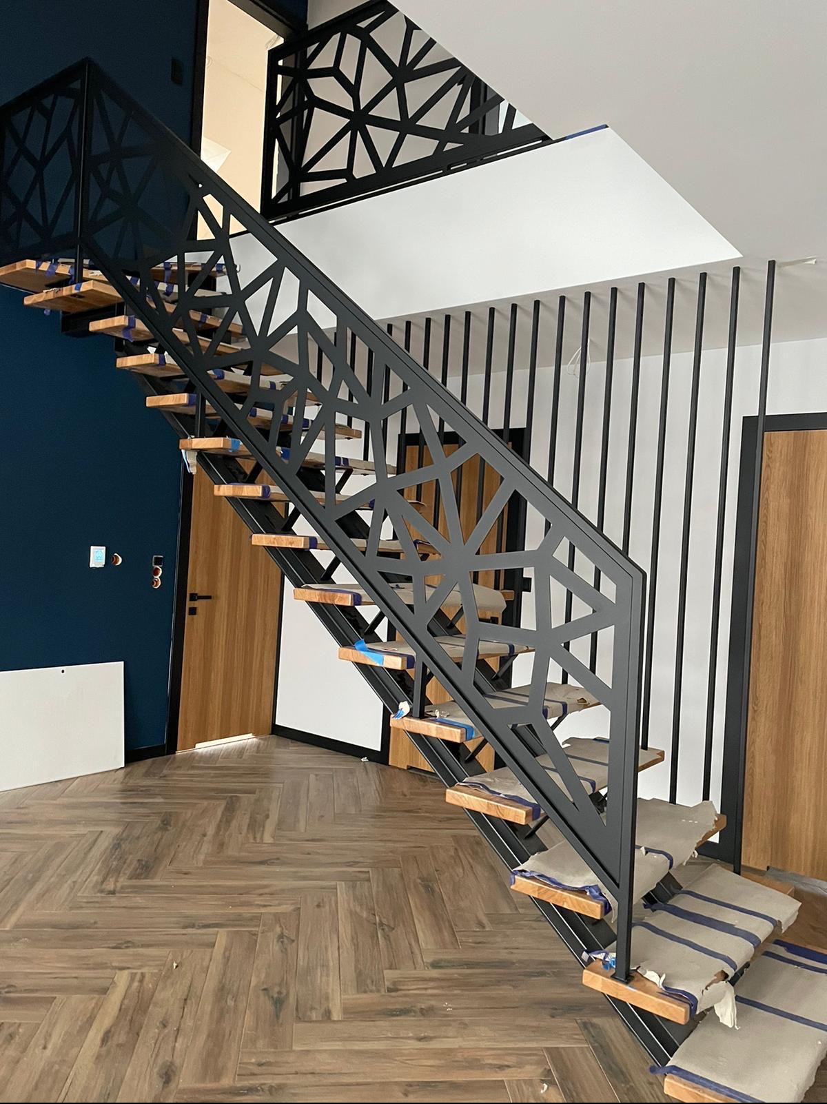 Nowoczesne schody z drewnianymi stopniami i metalową, geometryczną balustradą, zabezpieczone folią ochronną, na tle parkietu ułożonego w jodełkę i granatowej ściany.