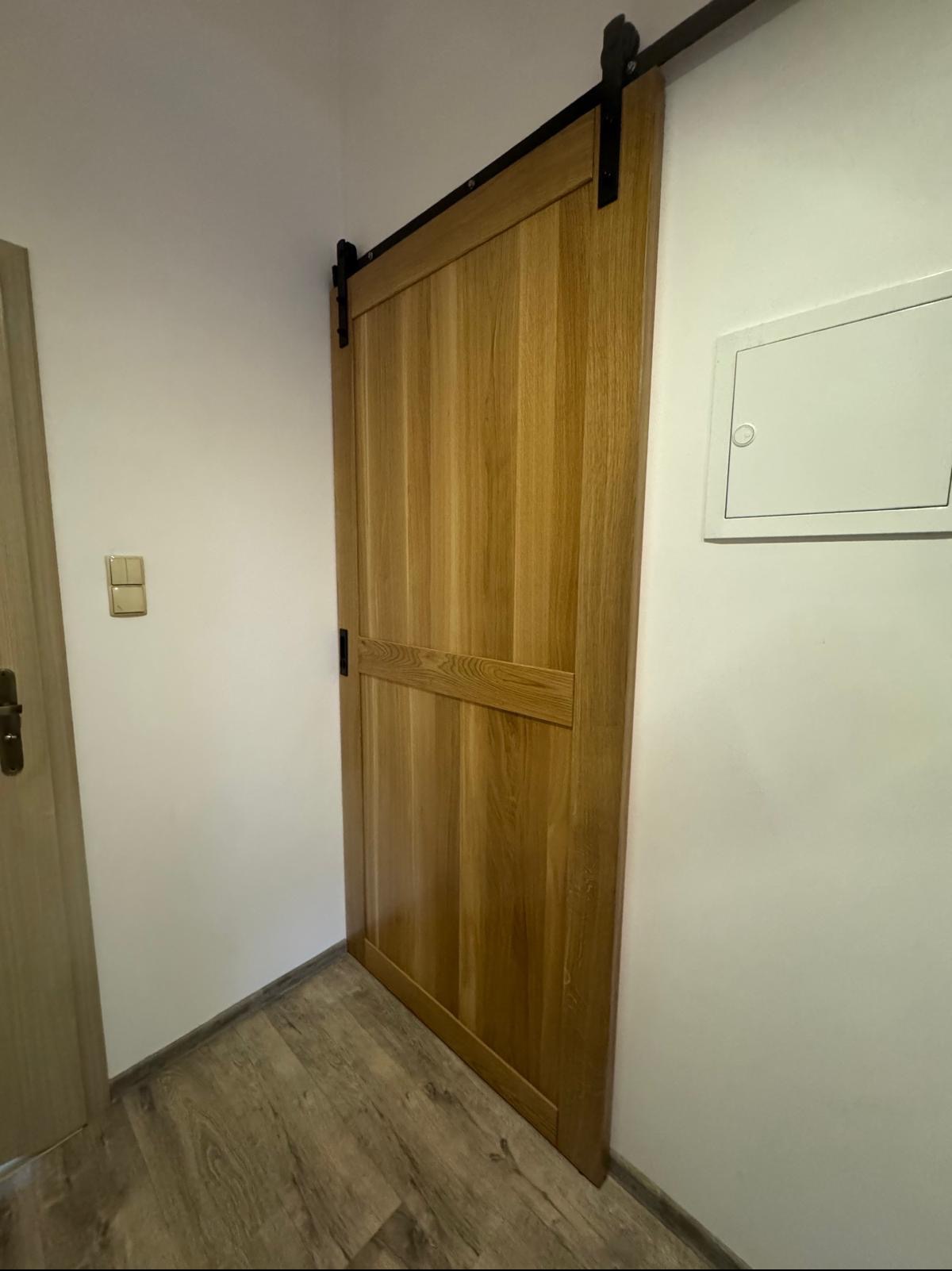 Suwane drzwi z jasnego drewna, zawieszone na czarnym, metalowym systemie szynowym, zamontowane w przejściu między pomieszczeniami, widoczne panele podłogowe imitujące drewno.