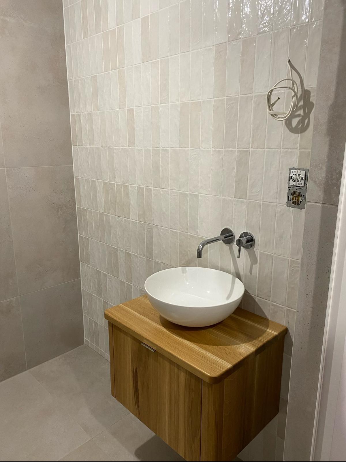 Biała, okrągła umywalka nablatowa na szafce z drewna w łazience z beżowymi, pionowo ułożonymi płytkami, widoczne gniazdka elektryczne w trakcie montażu.