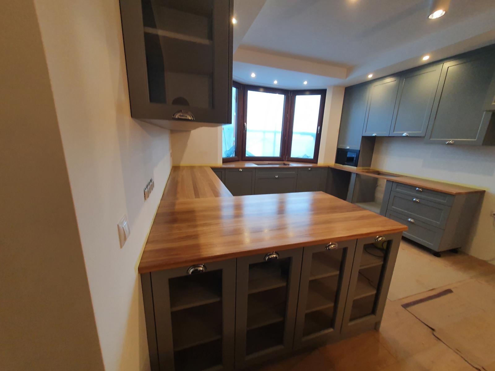 Szara zabudowa kuchenna z drewnianym blatem, widok na narożne okno i oświetlenie punktowe sufitowe, dolne szafki z przeszklonymi frontami.