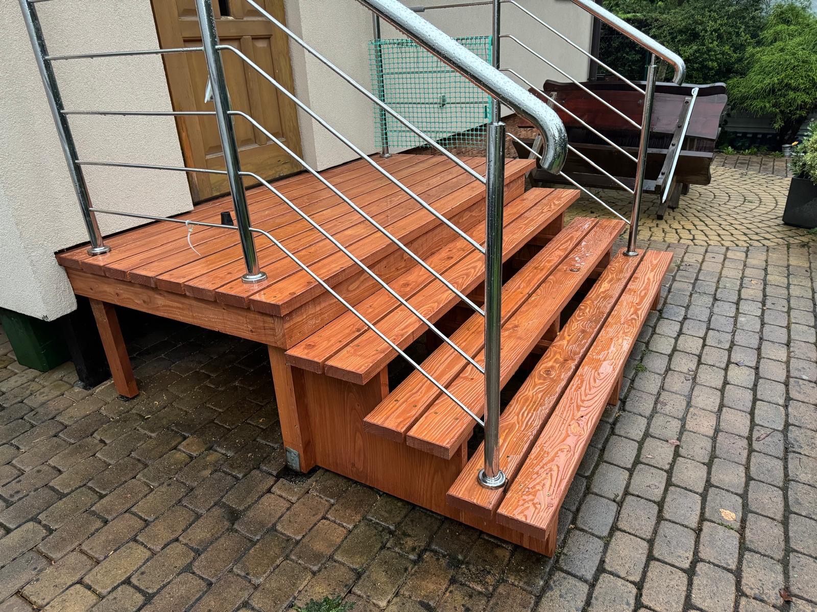 Zewnętrzne schody z drewna o ciepłym odcieniu, prowadzące do drzwi wejściowych, wyposażone w nowoczesną balustradę ze stali nierdzewnej, na mokrym brukowanym podłożu.