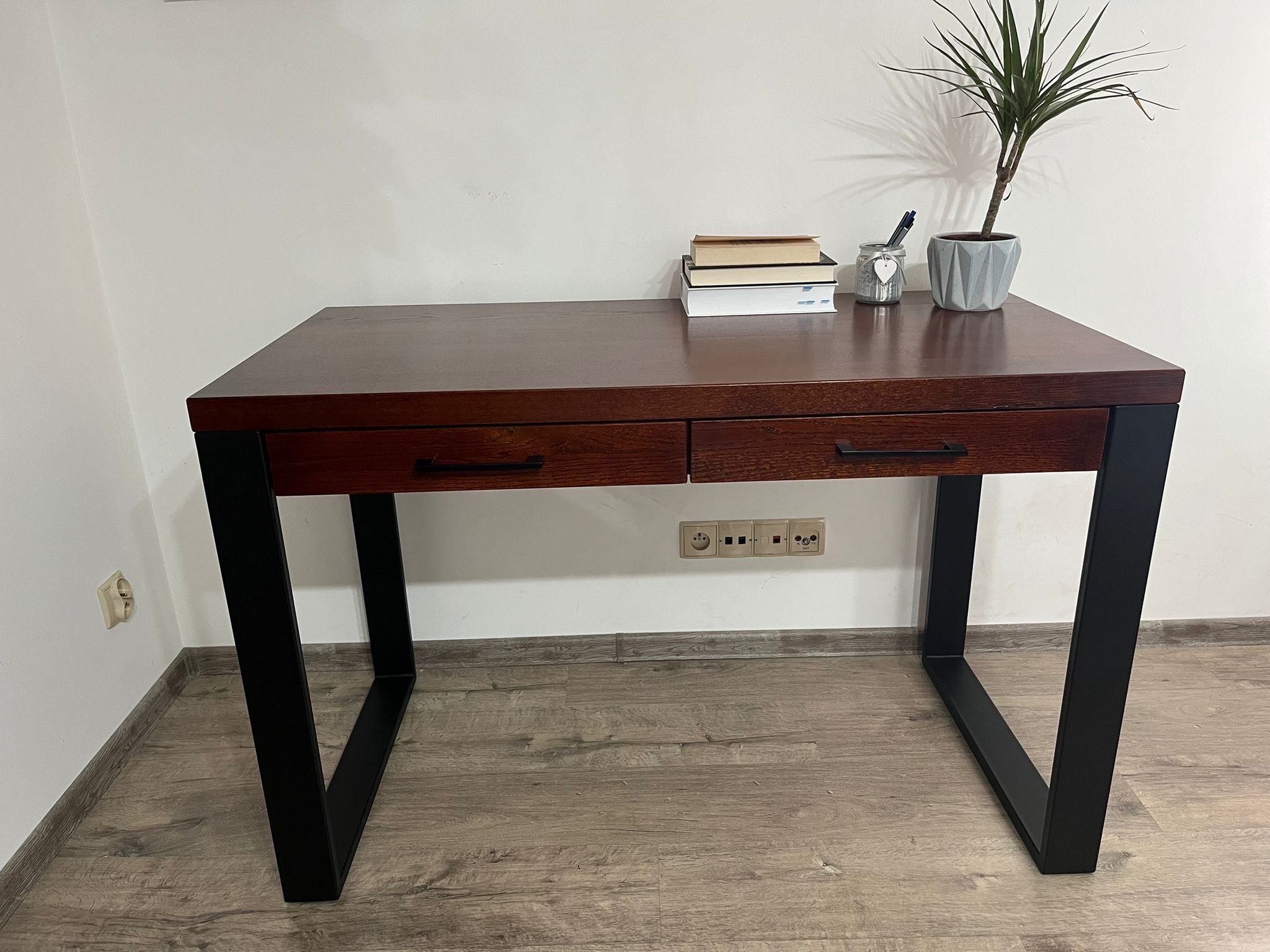 Biurko z ciemnego drewna z czarnymi, metalowymi nogami, z dwiema szufladami, na blacie książki, roślina doniczkowa i słoik z długopisami, na tle białej ściany z gniazdkami.