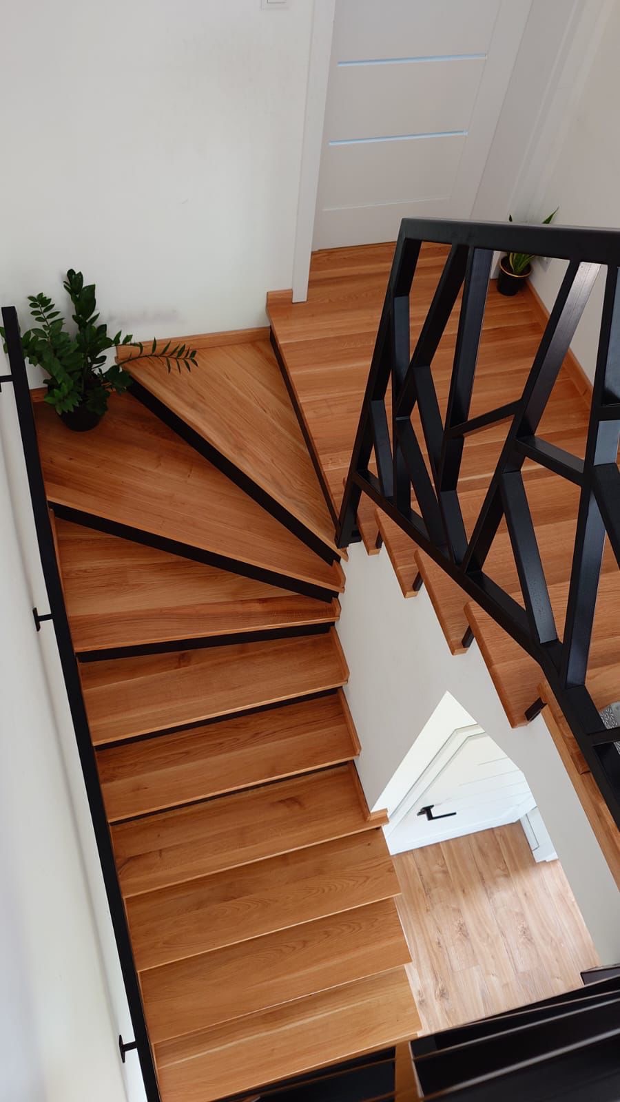 Drewniane schody z czarną, geometryczną balustradą, widok z góry, z rośliną doniczkową na pierwszym stopniu i widocznym fragmentem białych drzwi w tle.