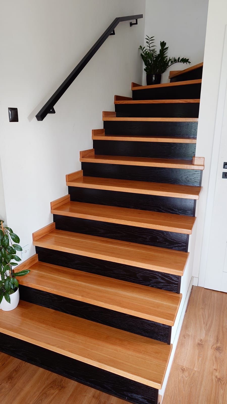 Nowoczesne schody z drewnianymi stopniami w odcieniu jasnego dębu i czarnymi podstopnicami, z minimalistyczną czarną balustradą i roślinami doniczkowymi na górze i dole biegu.