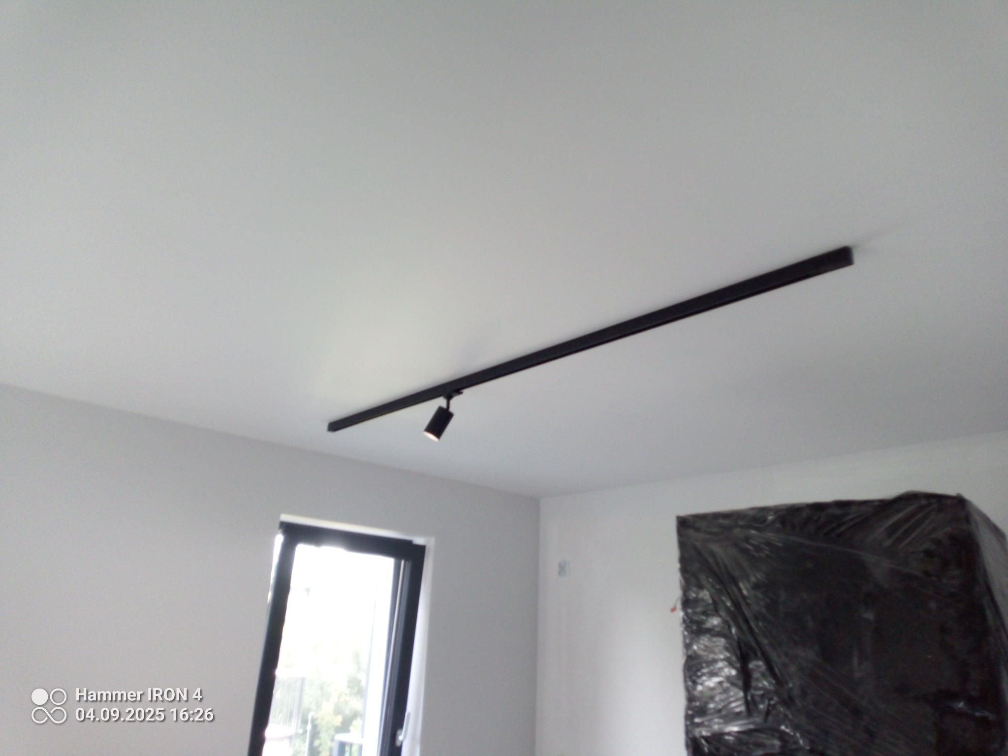 Nowoczesne oświetlenie szynowe z pojedynczym reflektorem, zamontowane na białym suficie w świeżo wykończonym wnętrzu. Okno i zabezpieczony mebel w tle.