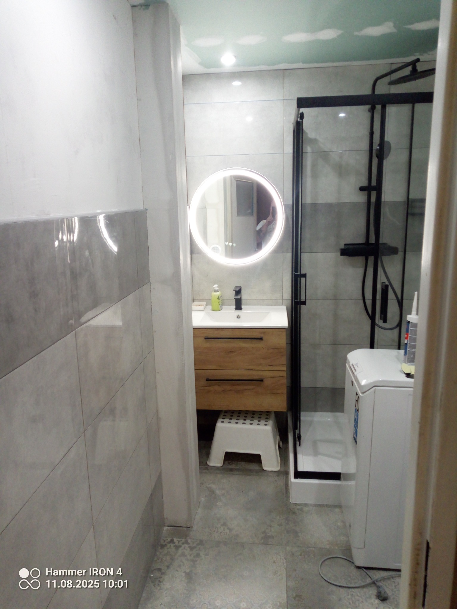 Nowoczesna łazienka po remoncie: szare płytki, okrągłe lustro z podświetleniem, drewniana szafka pod umywalką, czarna kabina prysznicowa i pralka. Widok z perspektywy drzwi.