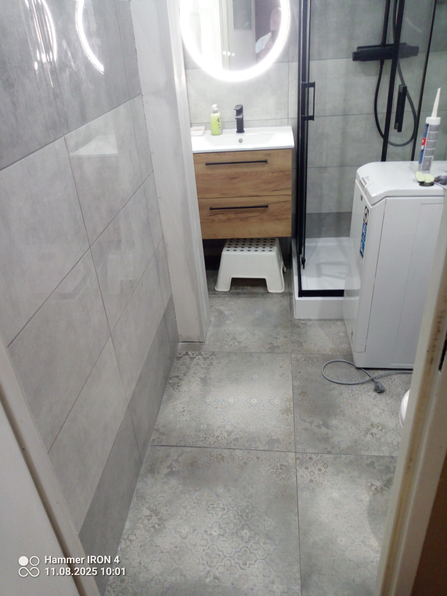 Wykończona łazienka z szarymi płytkami, drewnianą szafką pod umywalką i okrągłym lustrem z podświetleniem LED. Widoczna kabina prysznicowa i pralka.