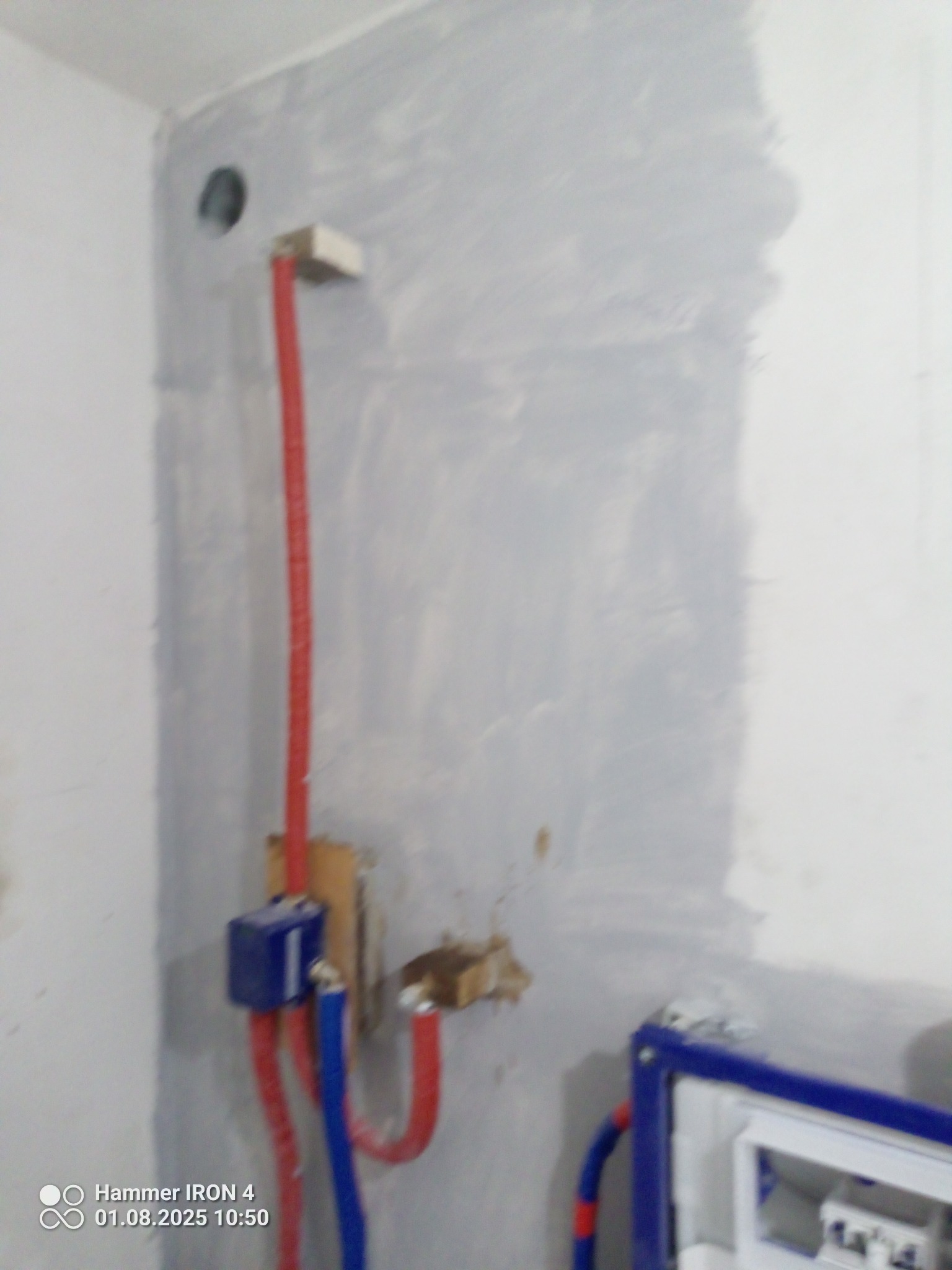 Ściana w trakcie remontu łazienki z widocznymi rurami instalacji wodnej w kolorze czerwonym i niebieskim, oraz elementami mocującymi. Świeżo położona gładź szpachlowa.