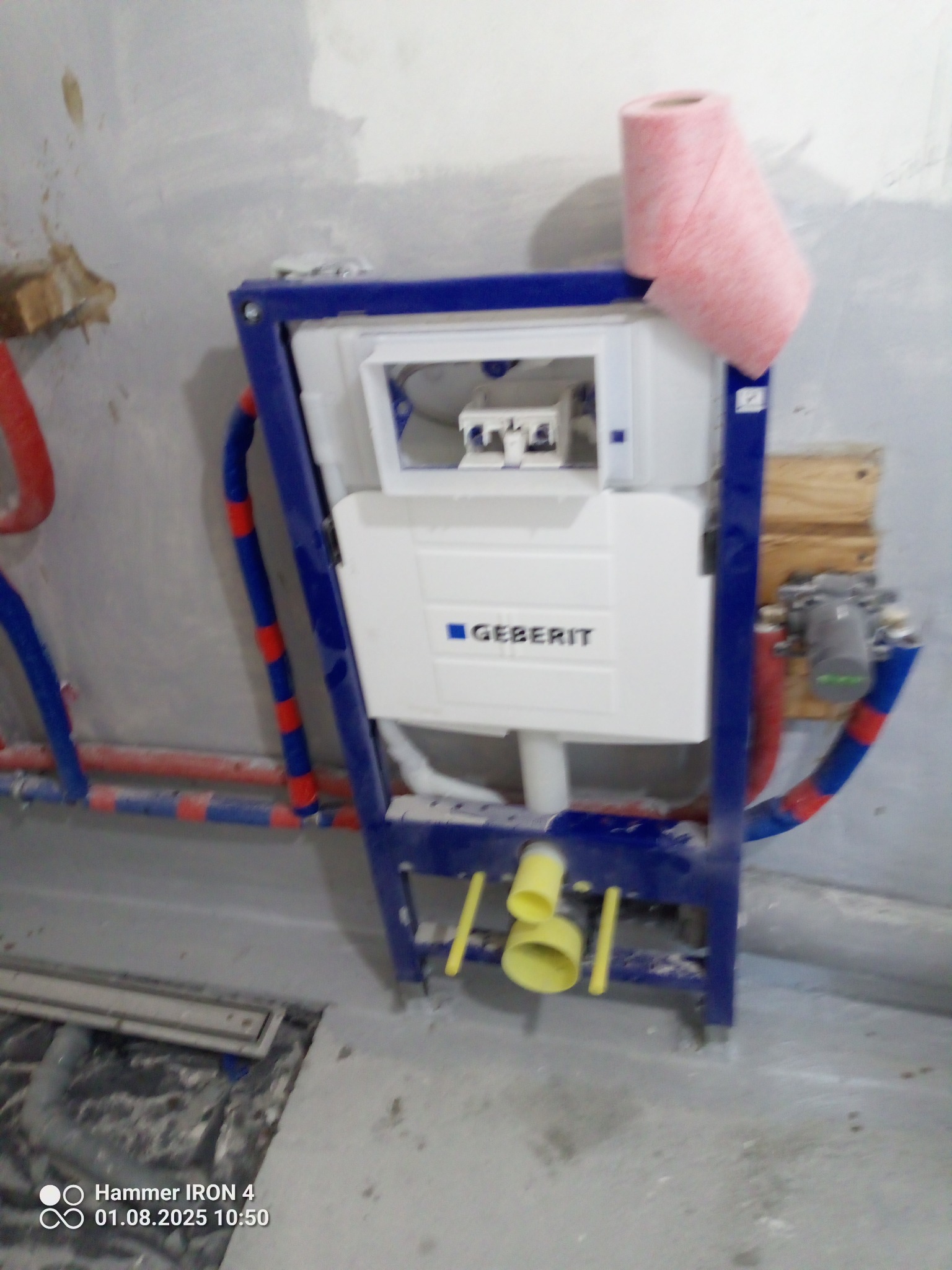 Stelaż podtynkowy Geberit z instalacją wodną w trakcie montażu w surowej łazience. Widoczne rury doprowadzające wodę i rolka różowego papieru na stelażu.