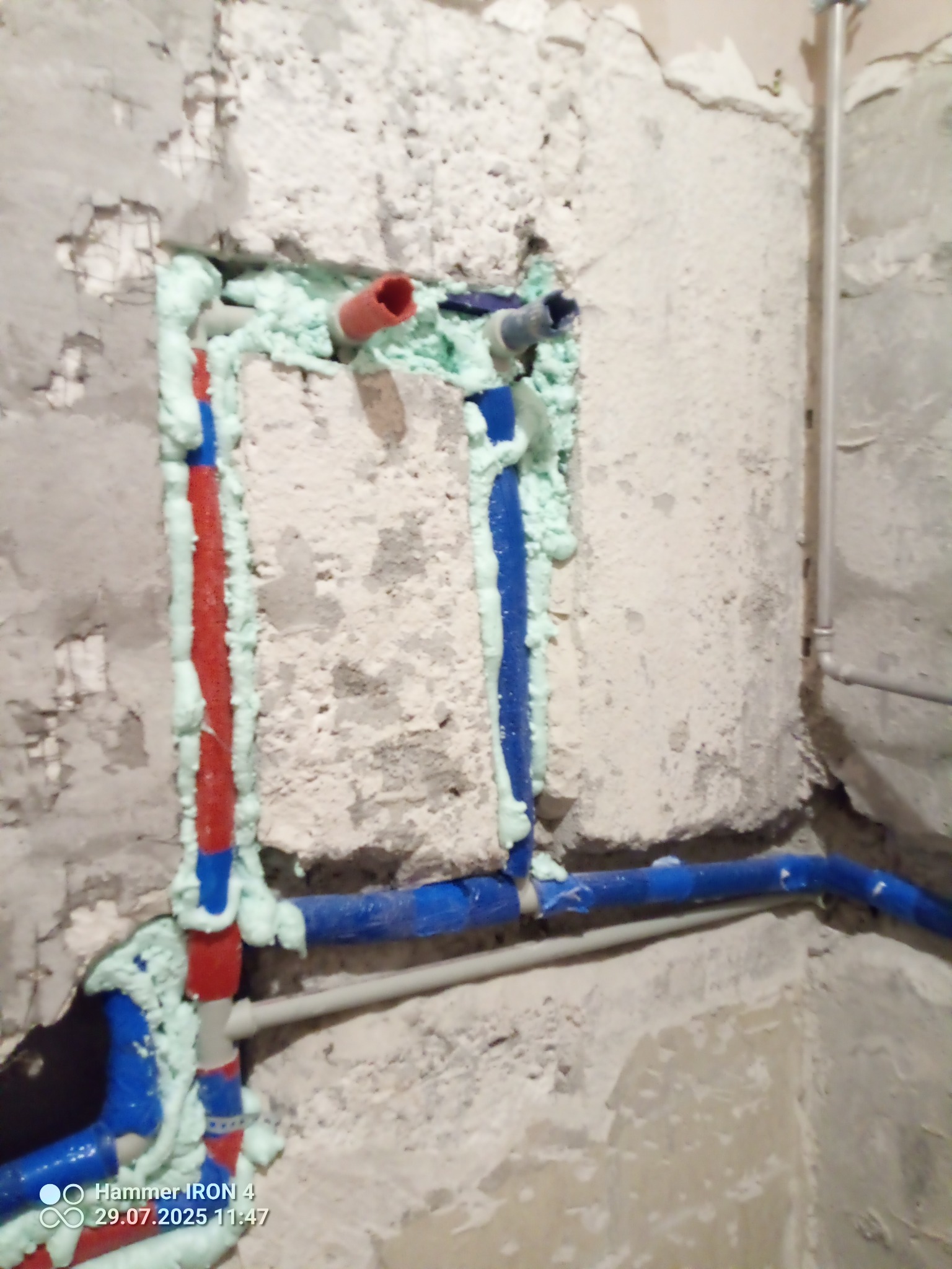 Wykute w ścianie rury wodne (czerwona i niebieska) zabezpieczone pianką montażową. Widoczne fragmenty ściany z surowym tynkiem.