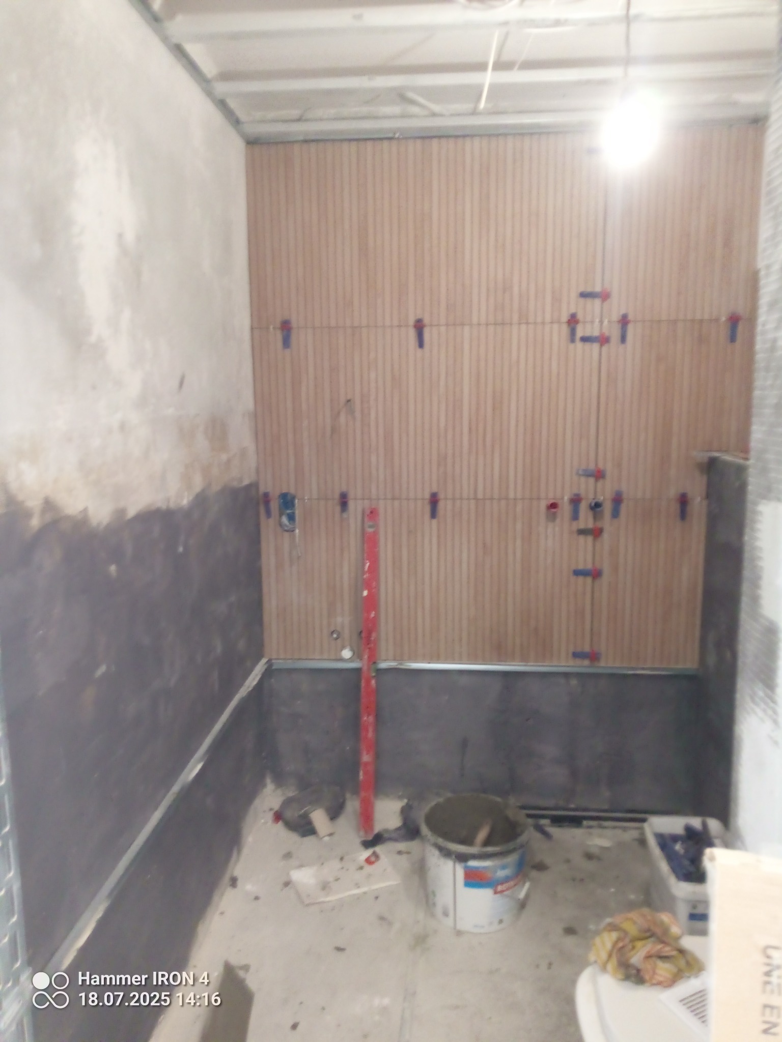 Montaż drewnopodobnych paneli ściennych w łazience, z użyciem krzyżyków dystansowych. Widoczny poziomica, wiadro z zaprawą i przygotowane podłoże.