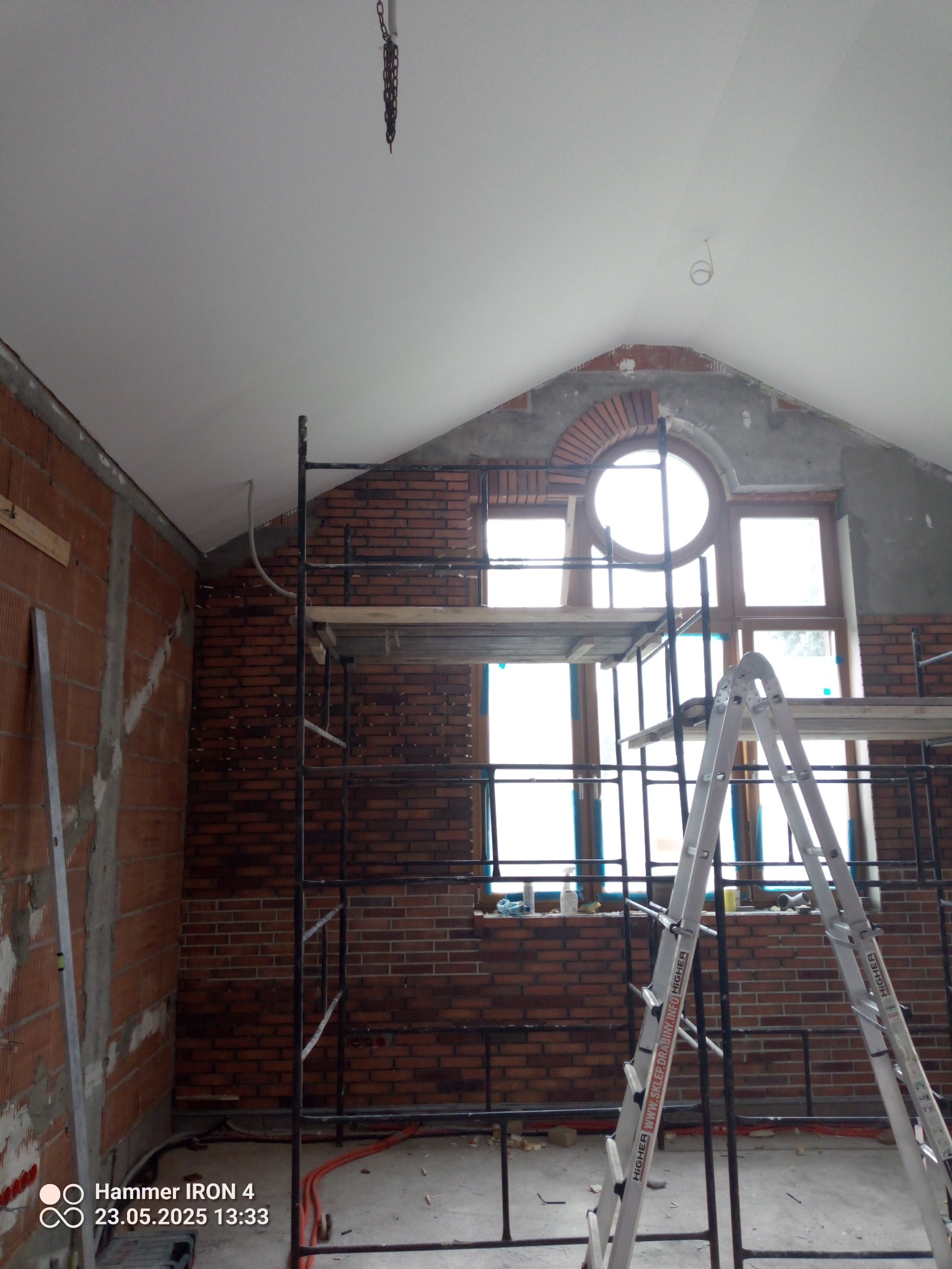 Wnętrze w trakcie prac murarskich z cegły klinkierowej, widoczne rusztowanie i drabina. Okrągłe okno w szczycie ściany. Częściowo otynkowana ściana.