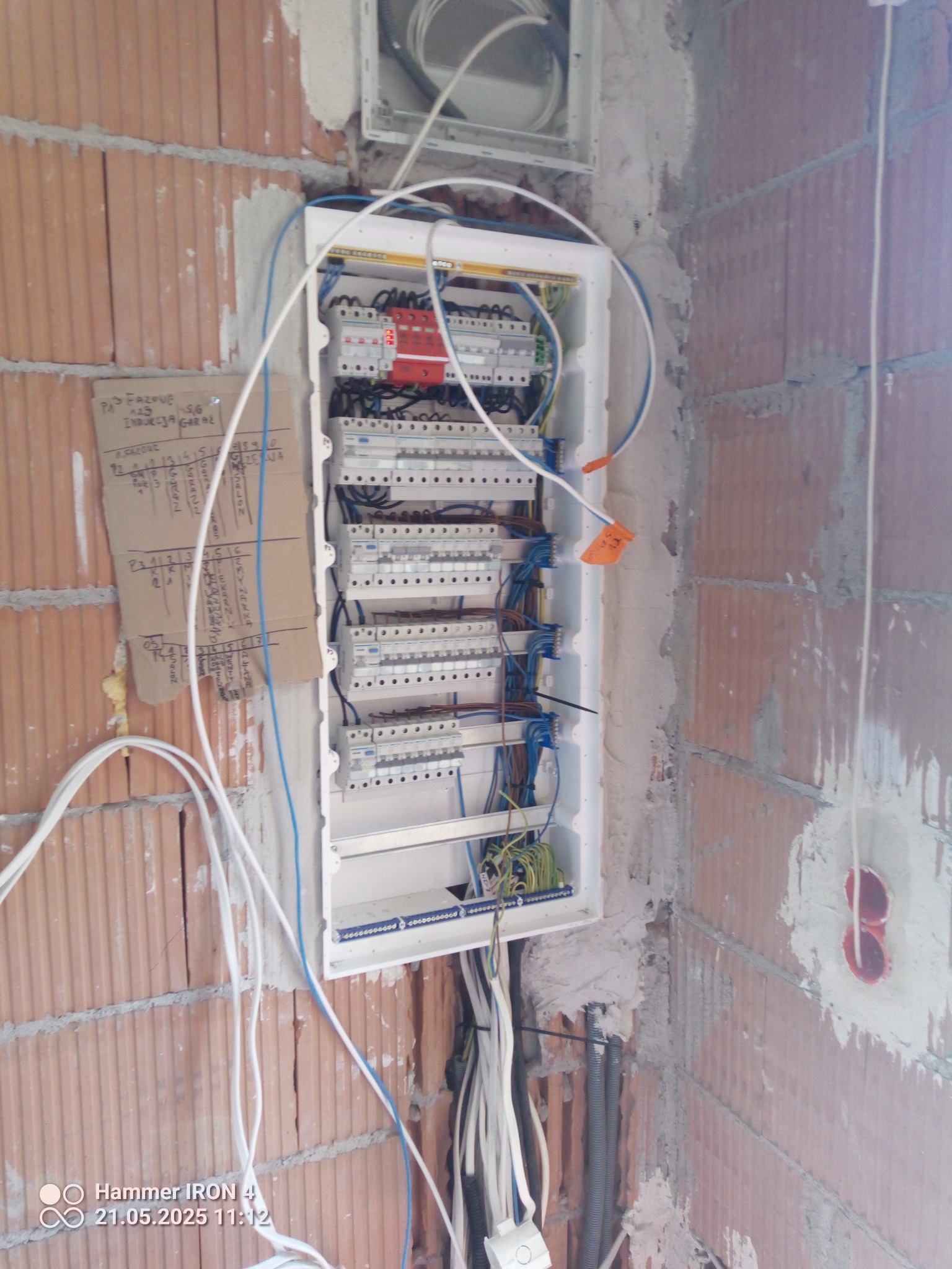 Rozdzielnia elektryczna z podłączonymi przewodami w surowym wnętrzu z cegły. Widoczne zabezpieczenia i kable. Na ścianie notatki odręczne.