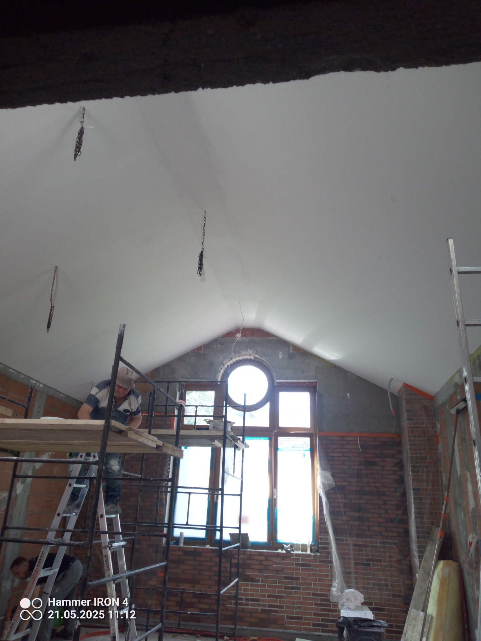 Montaż sufitu napinanego w pomieszczeniu z oknem o okrągłym kształcie. Widoczne rusztowania i pracownik podczas instalacji. Sufit biały, matowy.