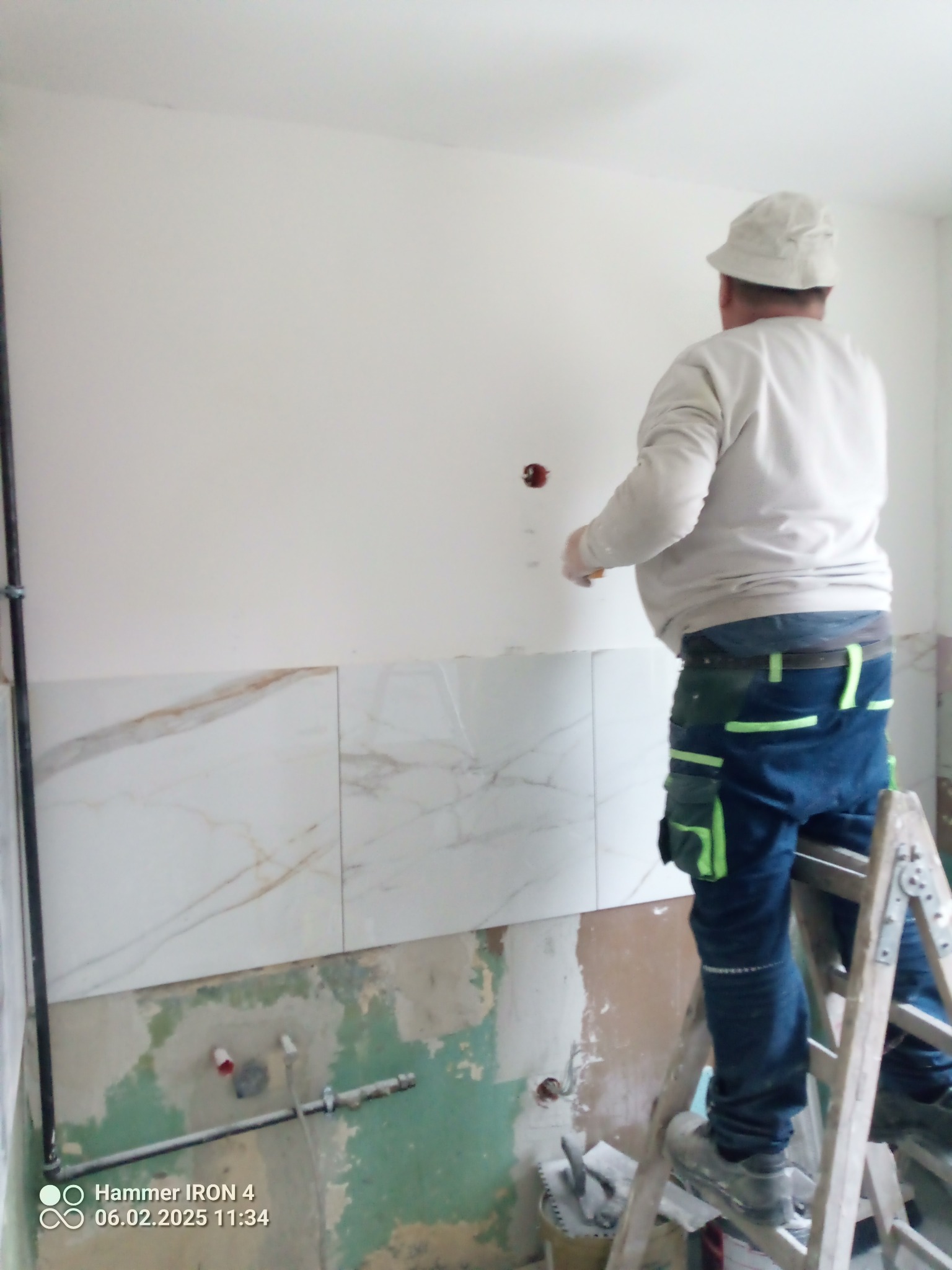Pracownik na drabinie układa marmurowe płytki na ścianie w łazience. Widoczne fragmenty instalacji i przygotowane podłoże. Praca w toku.