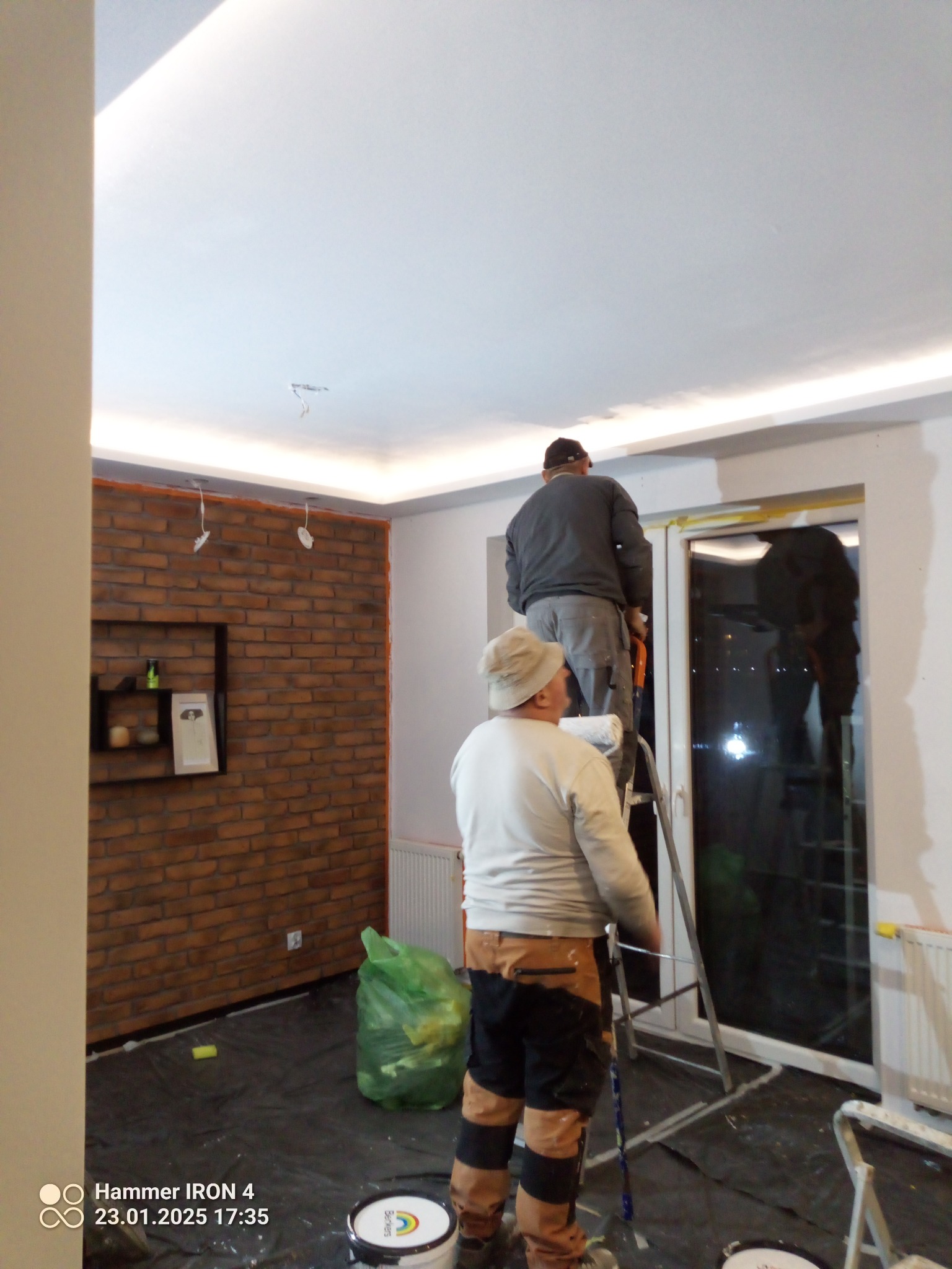 Dwóch malarzy na drabinie maluje sufit w pokoju z ceglaną ścianą. Podłoga zabezpieczona folią, widoczne puszki z farbą. Oświetlenie sufitowe LED.