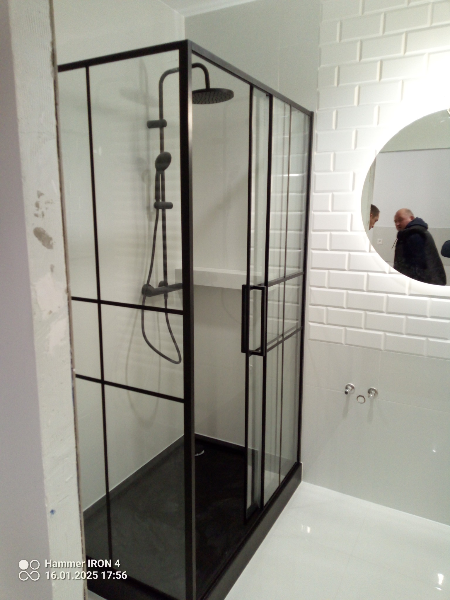 Nowoczesna kabina prysznicowa z czarnymi profilami i szklanymi ściankami, białe płytki na ścianach, okrągłe lustro z odbiciem osoby. Minimalistyczny design.