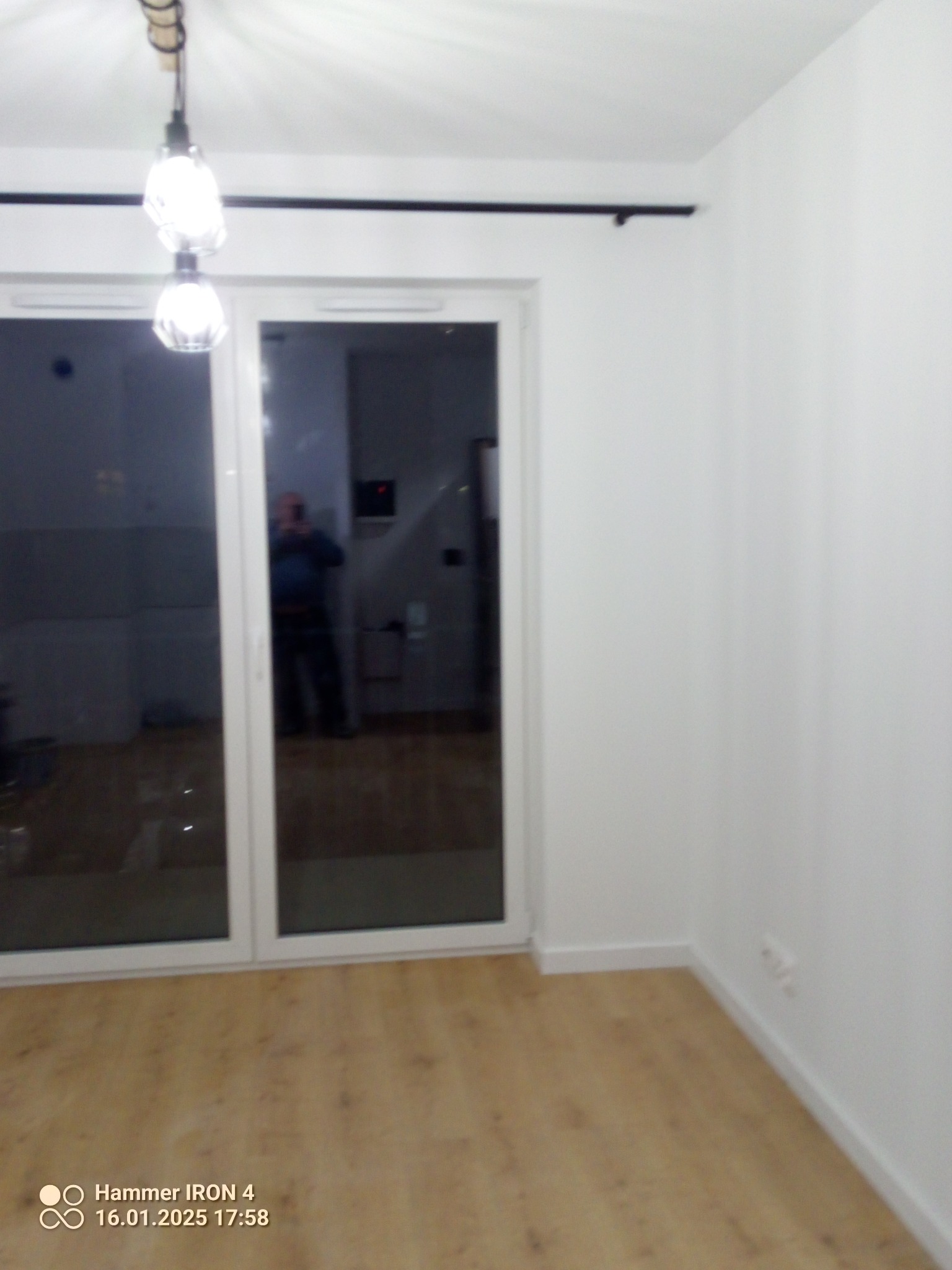 Jasne wnętrze po remoncie: parkiet, białe ściany, okno balkonowe z czarną zasłoną. Widoczny fragment lampy sufitowej. W oknie odbija się osoba robiąca zdjęcie.