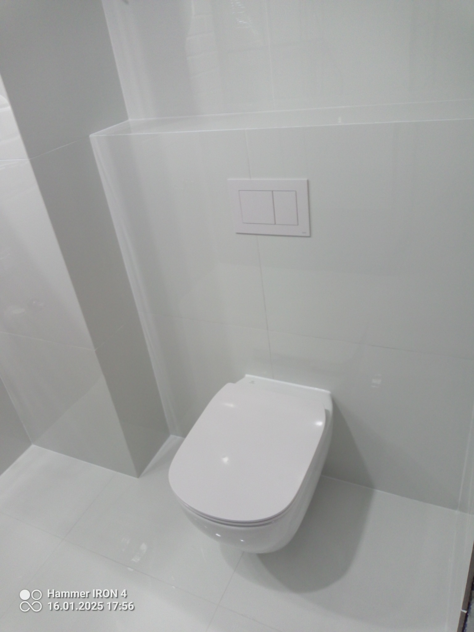 Biała, podwieszana toaleta w minimalistycznej łazience z błyszczącymi, białymi płytkami i ukrytym systemem spłukiwania. Nowoczesny design.