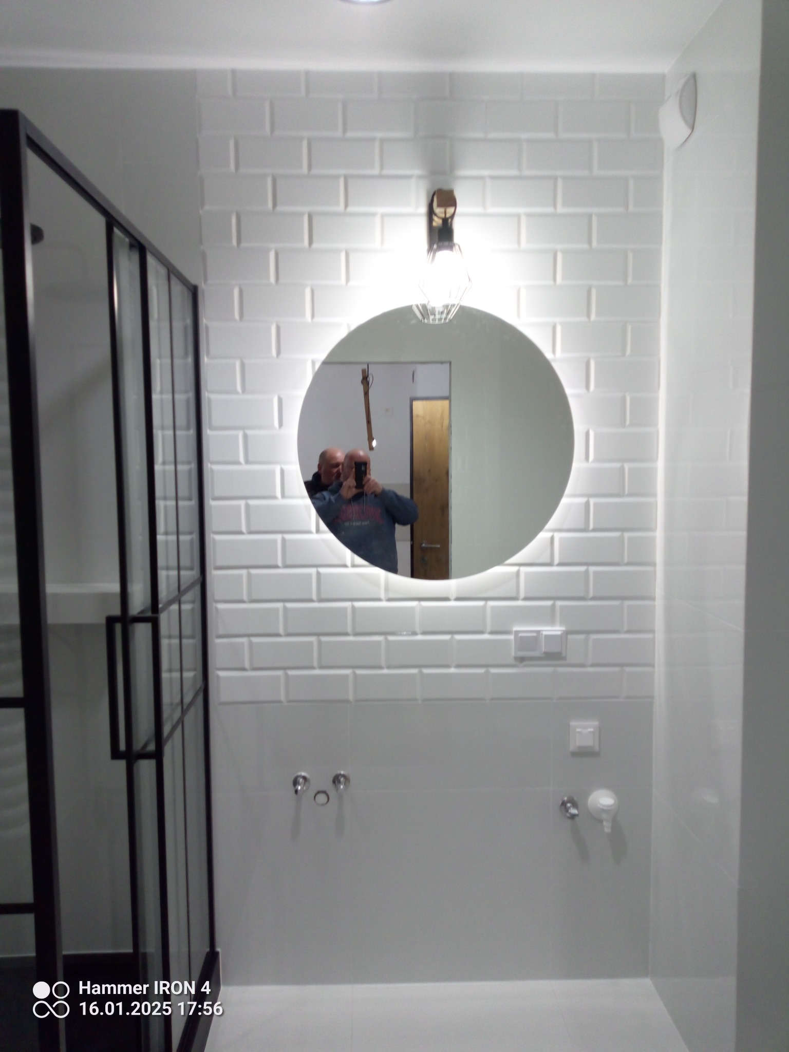 Biała łazienka z kabiną prysznicową typu walk-in i okrągłym lustrem z podświetleniem. Ściany wyłożone białymi, prostokątnymi płytkami imitującymi cegłę.