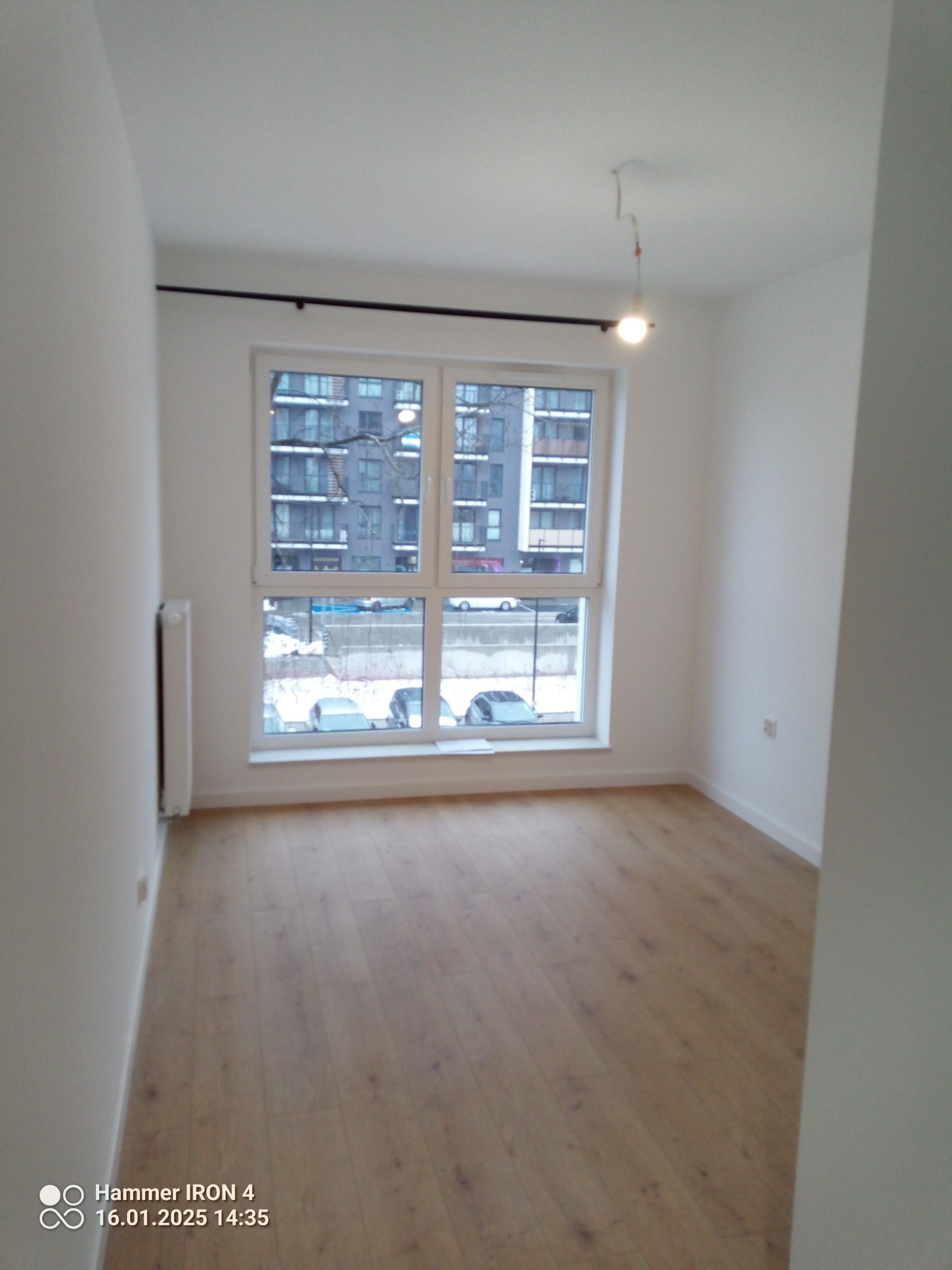 Jasne, puste wnętrze pokoju z drewnianą podłogą i dużym oknem z widokiem na budynek mieszkalny w zimowej scenerii. Widoczny grzejnik i niedokończone oświetlenie sufitowe.