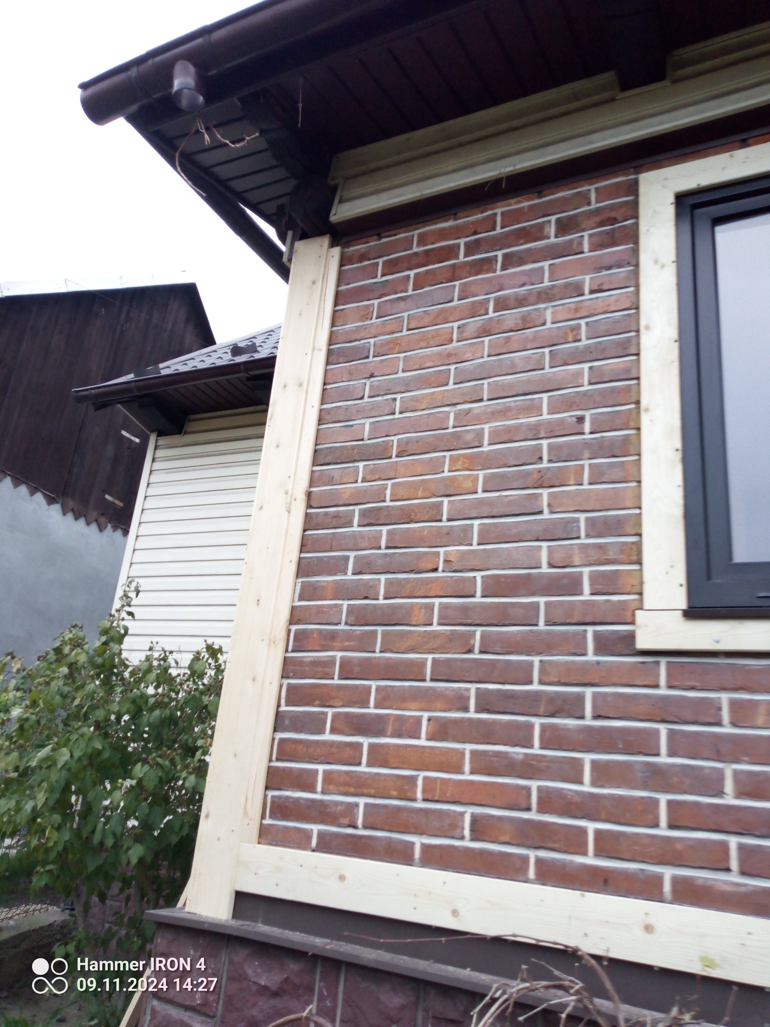 Fragment elewacji budynku z cegły klinkierowej, z drewnianą ramą okienną w trakcie montażu. Widoczny fragment dachu i okna. Ujęcie z dołu, podkreślające detale wykończenia.