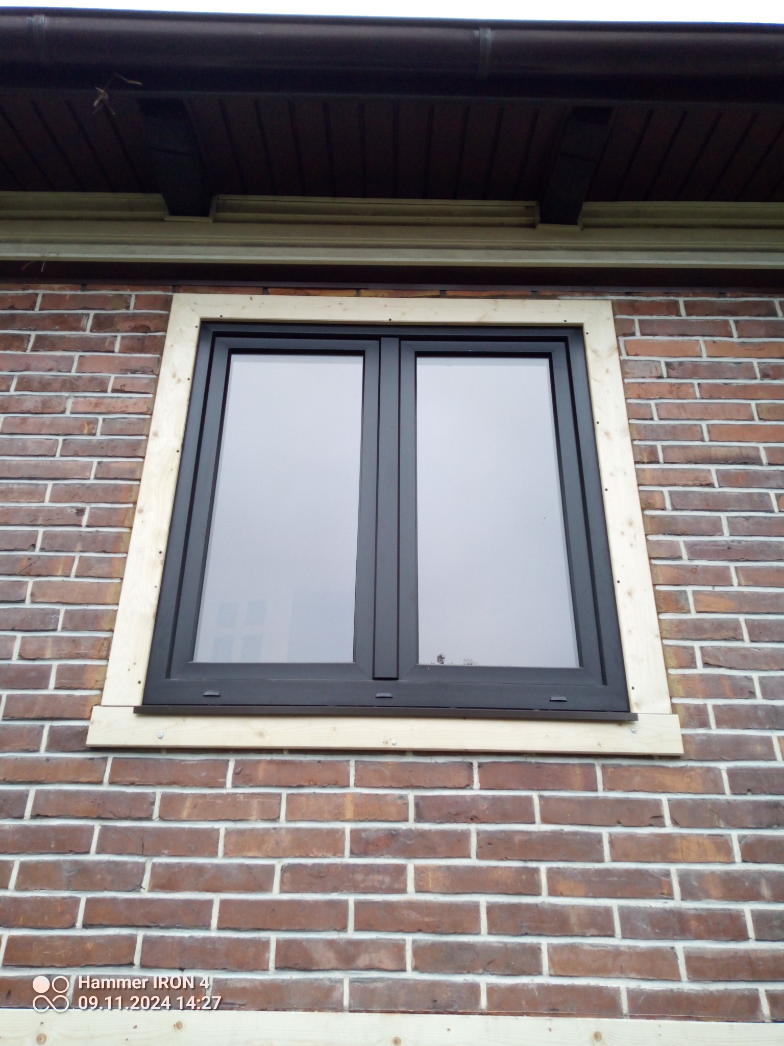 Ciemne okno PCV w surowej, drewnianej ramie montażowej na tle ceglanej ściany zewnętrznej budynku. Widok z dołu, fragment dachu widoczny u góry.