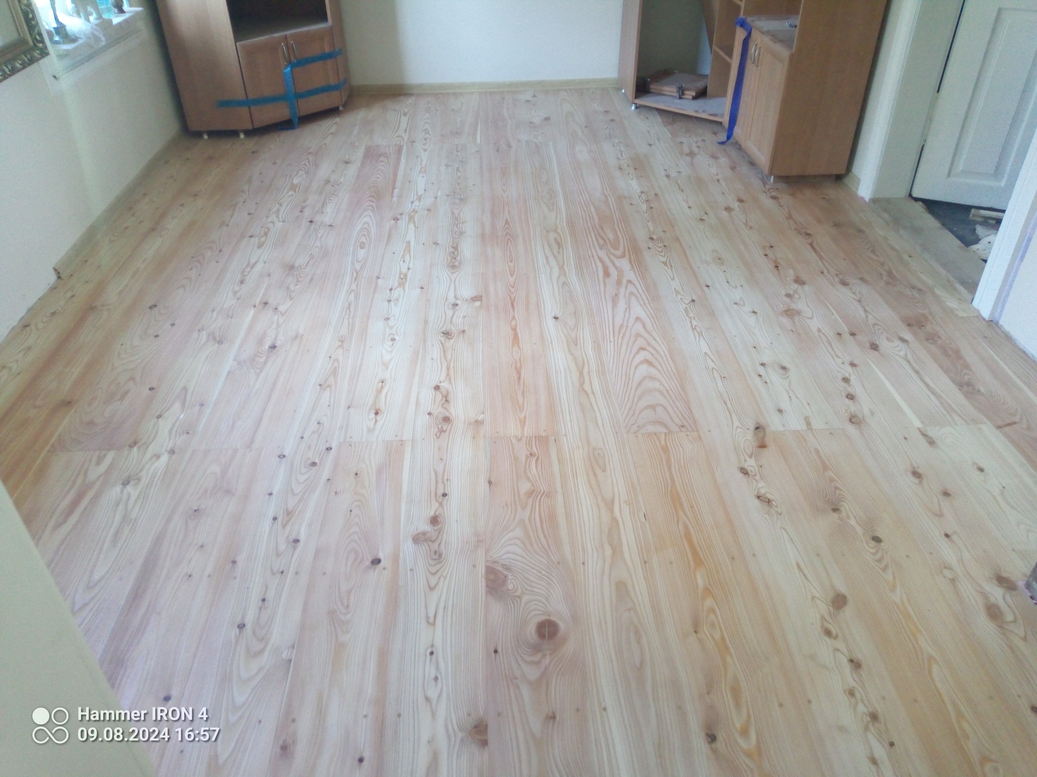 Jasna, drewniana podłoga z widocznym usłojeniem, świeżo po ułożeniu. W tle meble zabezpieczone taśmą malarską i fragment białych drzwi.